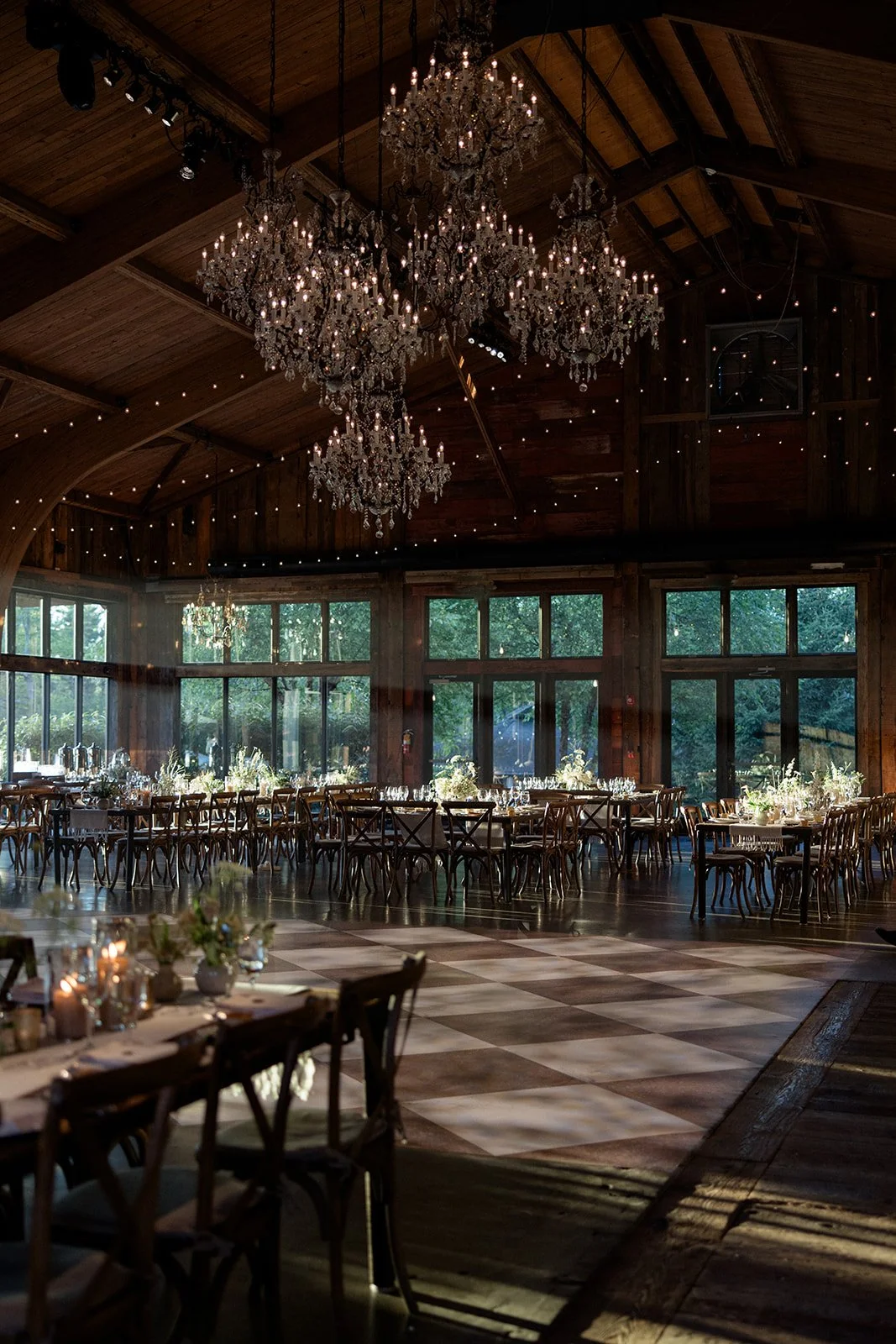 Elegant event hall with large crystal chandeliers hanging from wooden ceiling, decorated tables with floral centerpieces and glassware, and large windows letting in natural light.