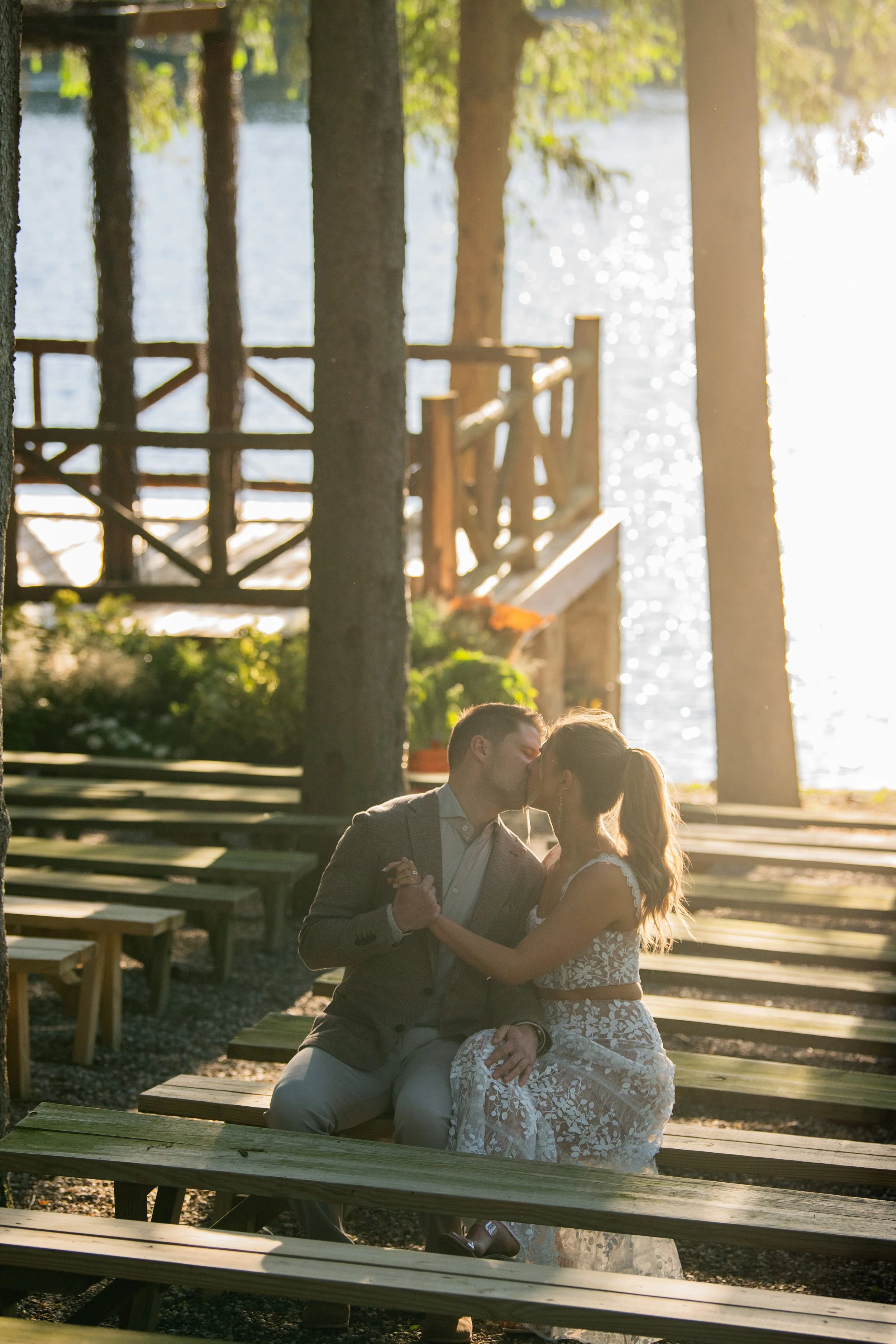 A couple dressed in wedding attire sitting on a bench and sharing a kiss near a lake, surrounded by wooden walkways and trees.