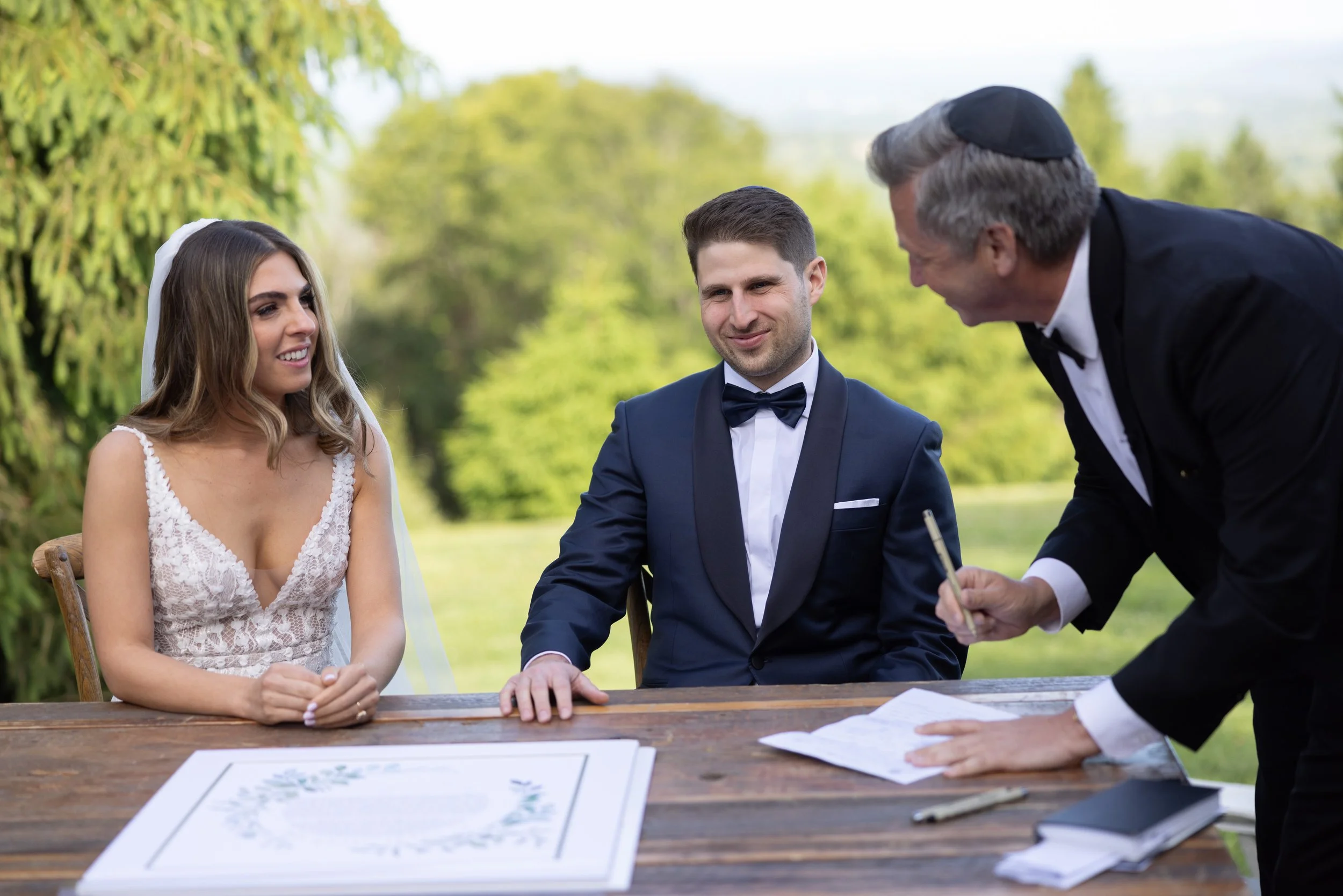 A couple getting married outdoors, sitting at a wooden table with a religious officiant.