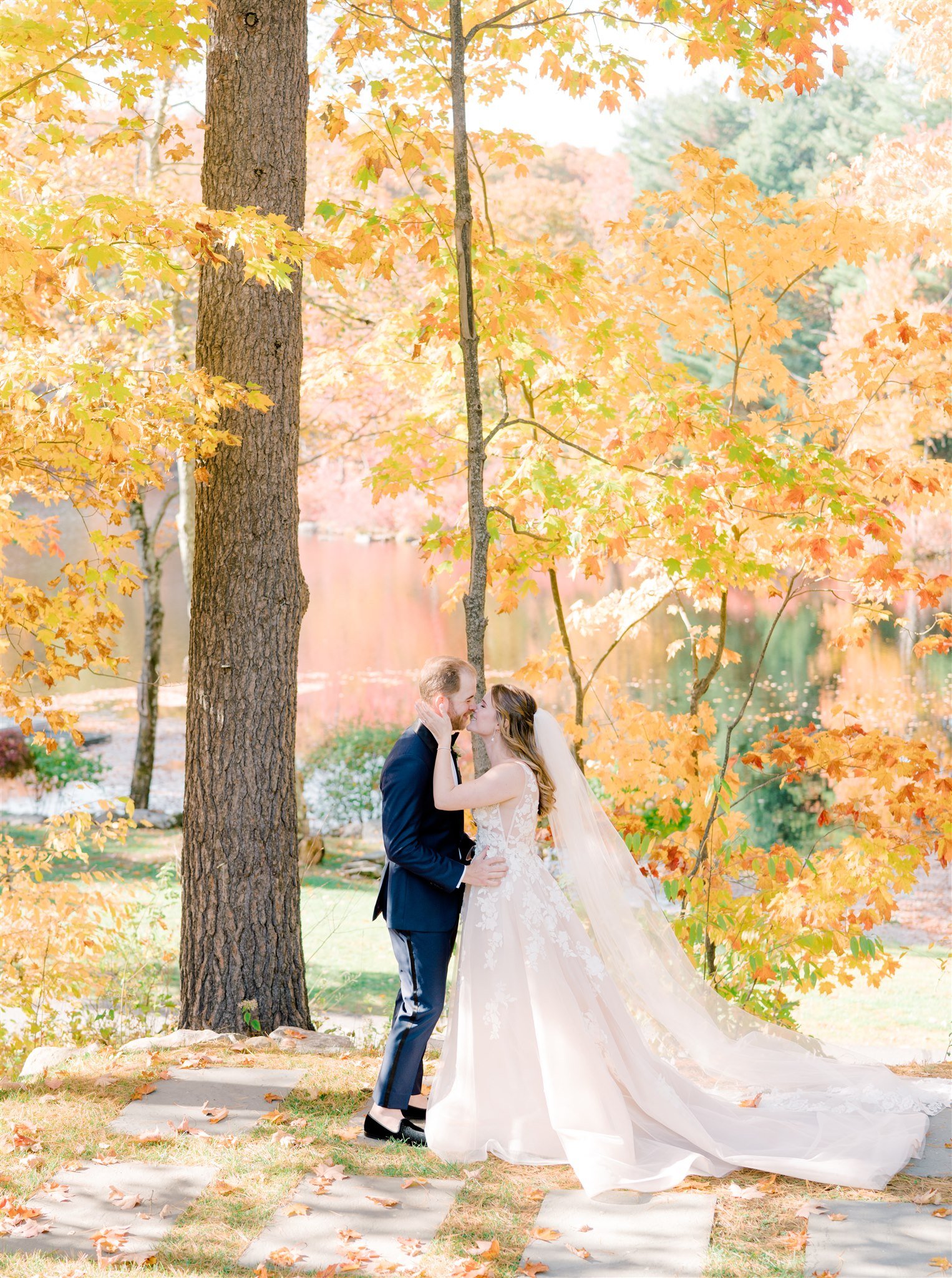 A bride and groom in wedding attire sharing a kiss outdoors in a park with fall foliage and trees with orange leaves.
