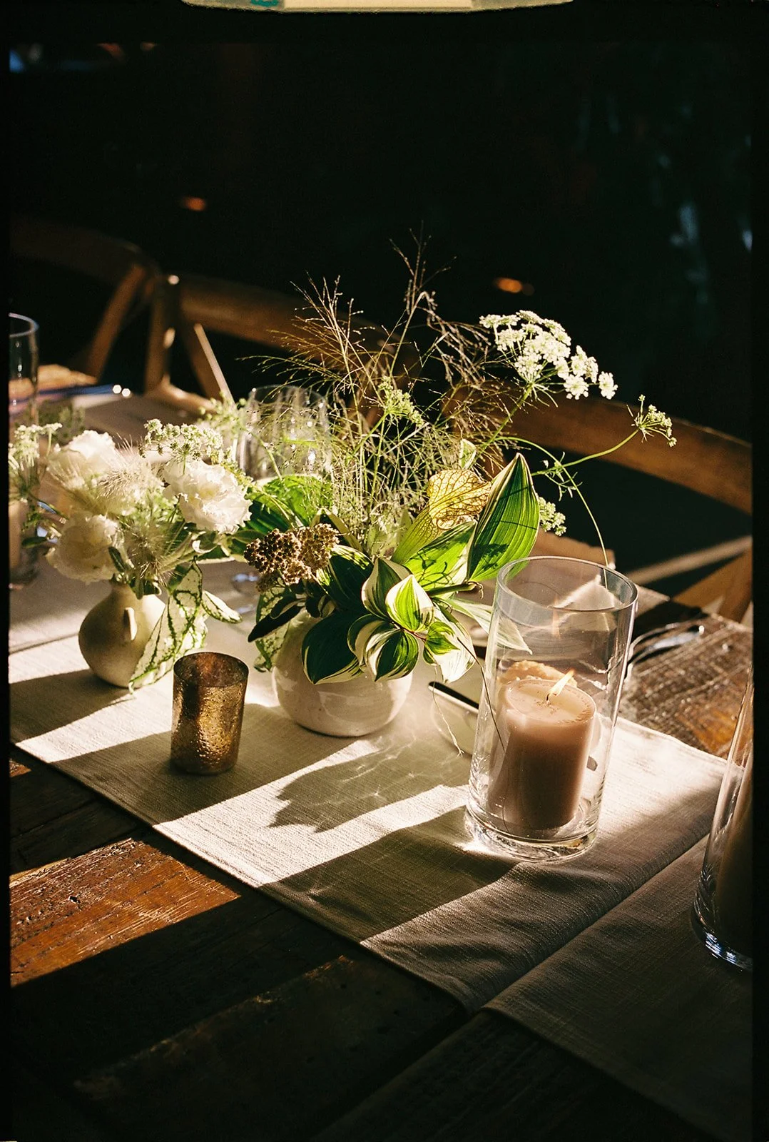 A rustic dining table with a beige table runner, decorated with a floral centerpiece of white flowers and green leaves, a lit white candle in a glass holder, and a small metallic candle holder, in a dimly lit setting with warm lighting and shadows.