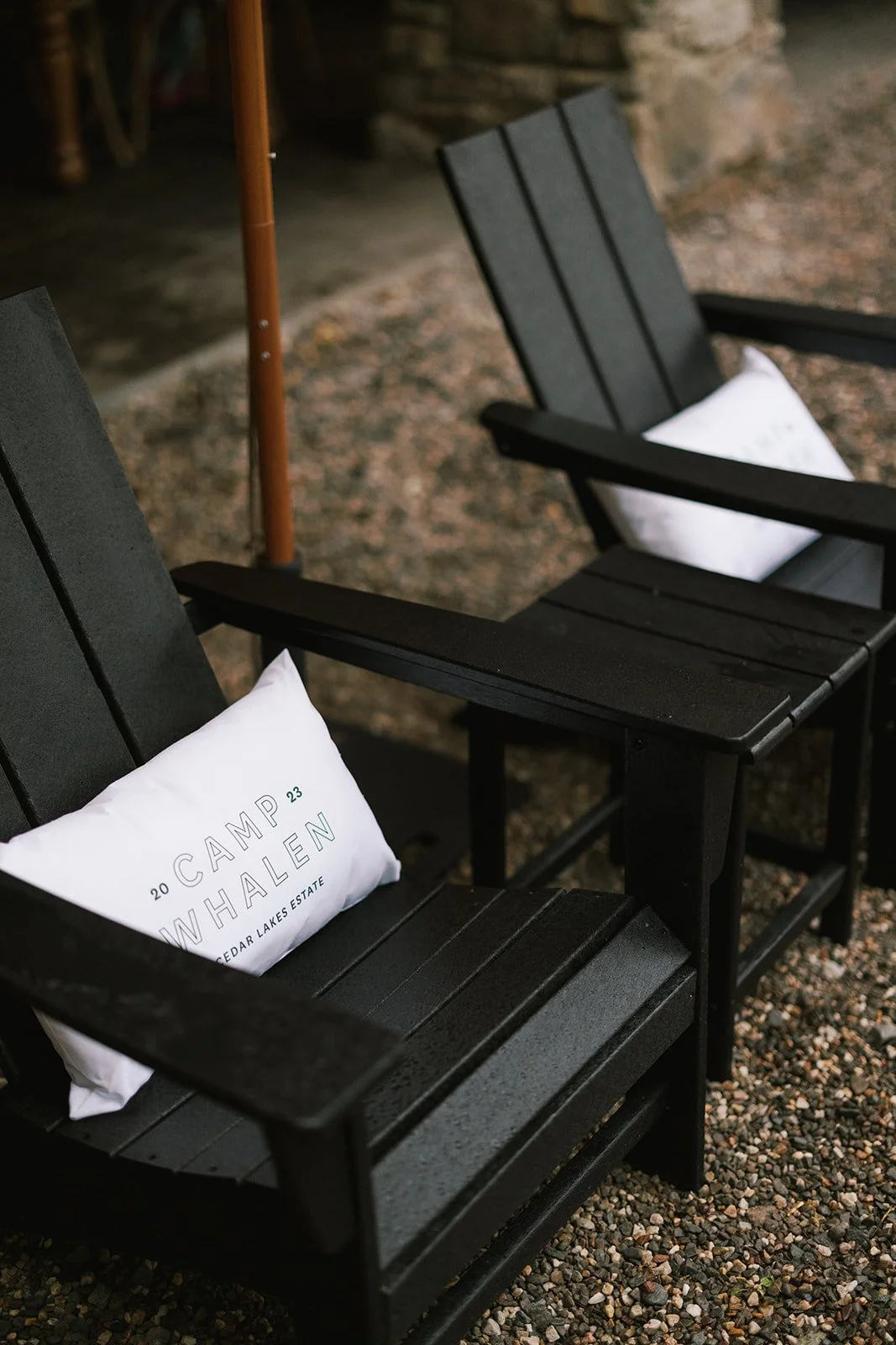 Two black Adirondack chairs with white pillows, one pillow reads "CAMP WHALE 2023 Cedar Lakes Estate," on a gravel surface. An orange patio umbrella pole is in the background.