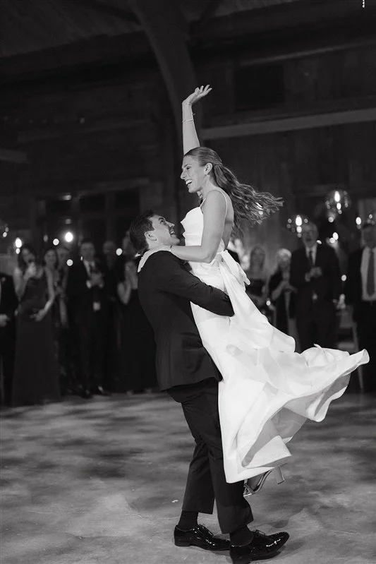 A couple dancing at a formal event with guests clapping in the background. The man is lifting the woman while spinning her, and they are dressed in formal attire.