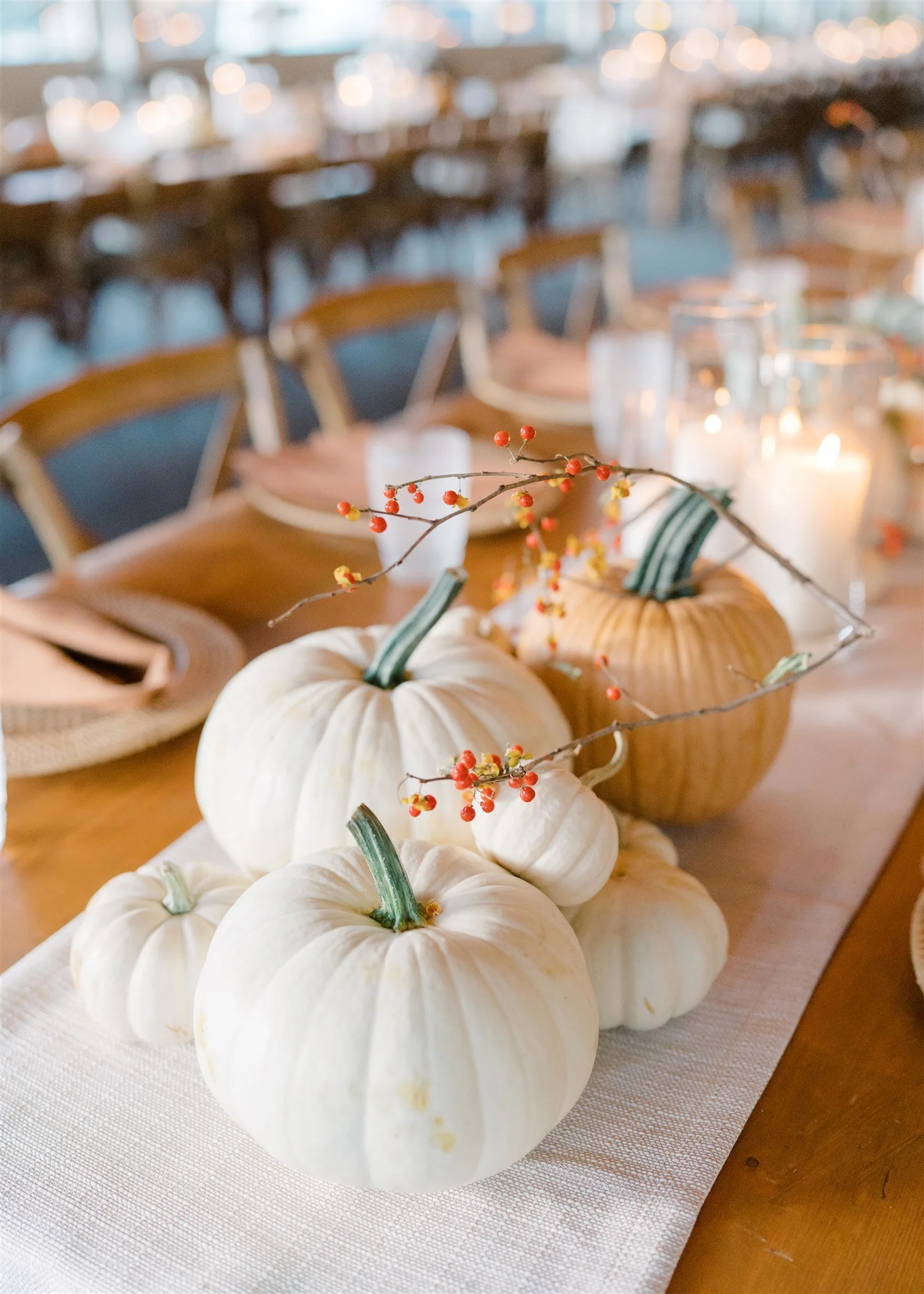 A table decorated with white and orange pumpkins, a branch with red berries, candles, glasses, and napkins, set for a fall or Thanksgiving meal.