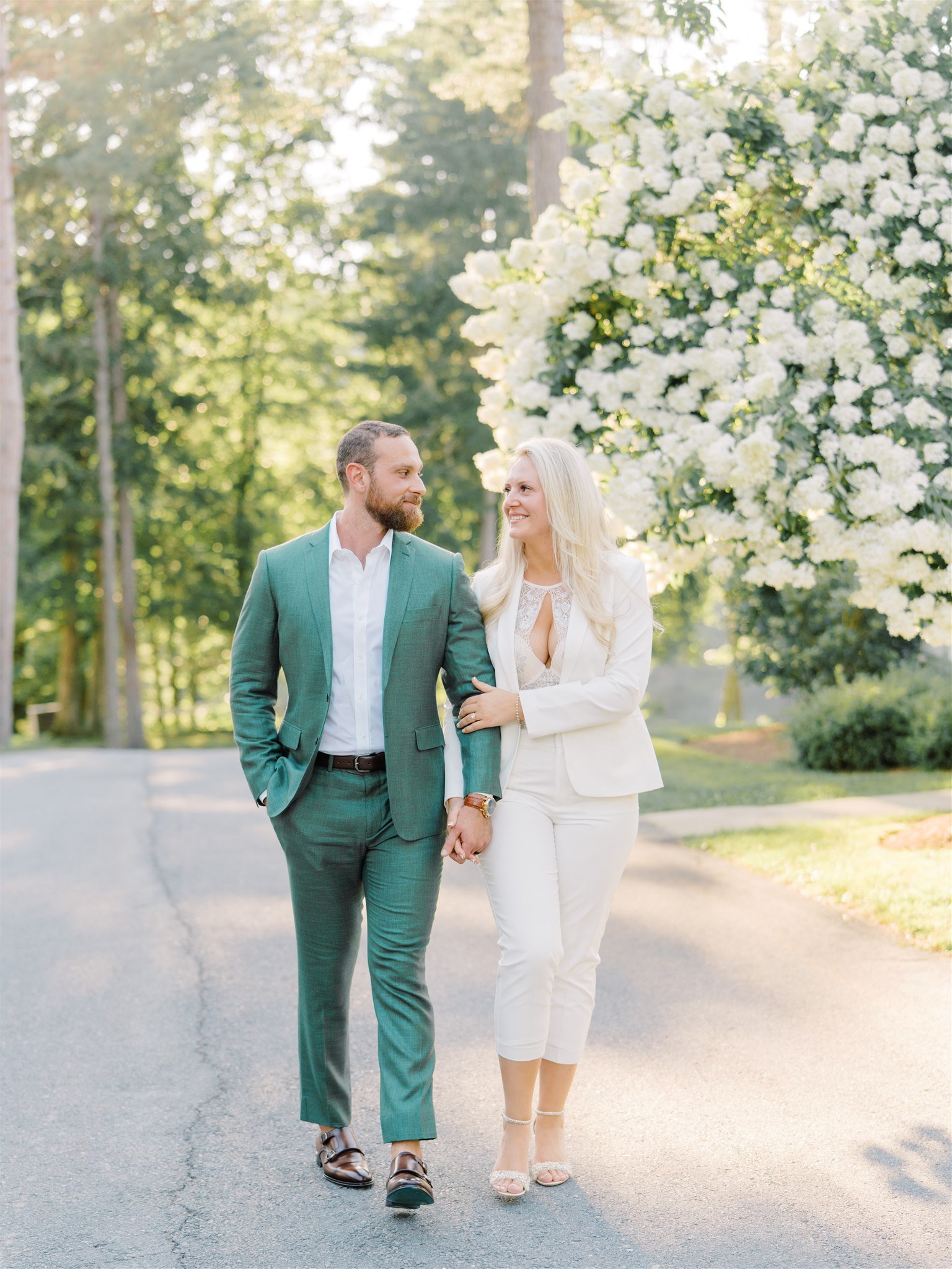 A couple walking arm-in-arm outdoors near blooming white flowers and tall trees, enjoying a sunny day.