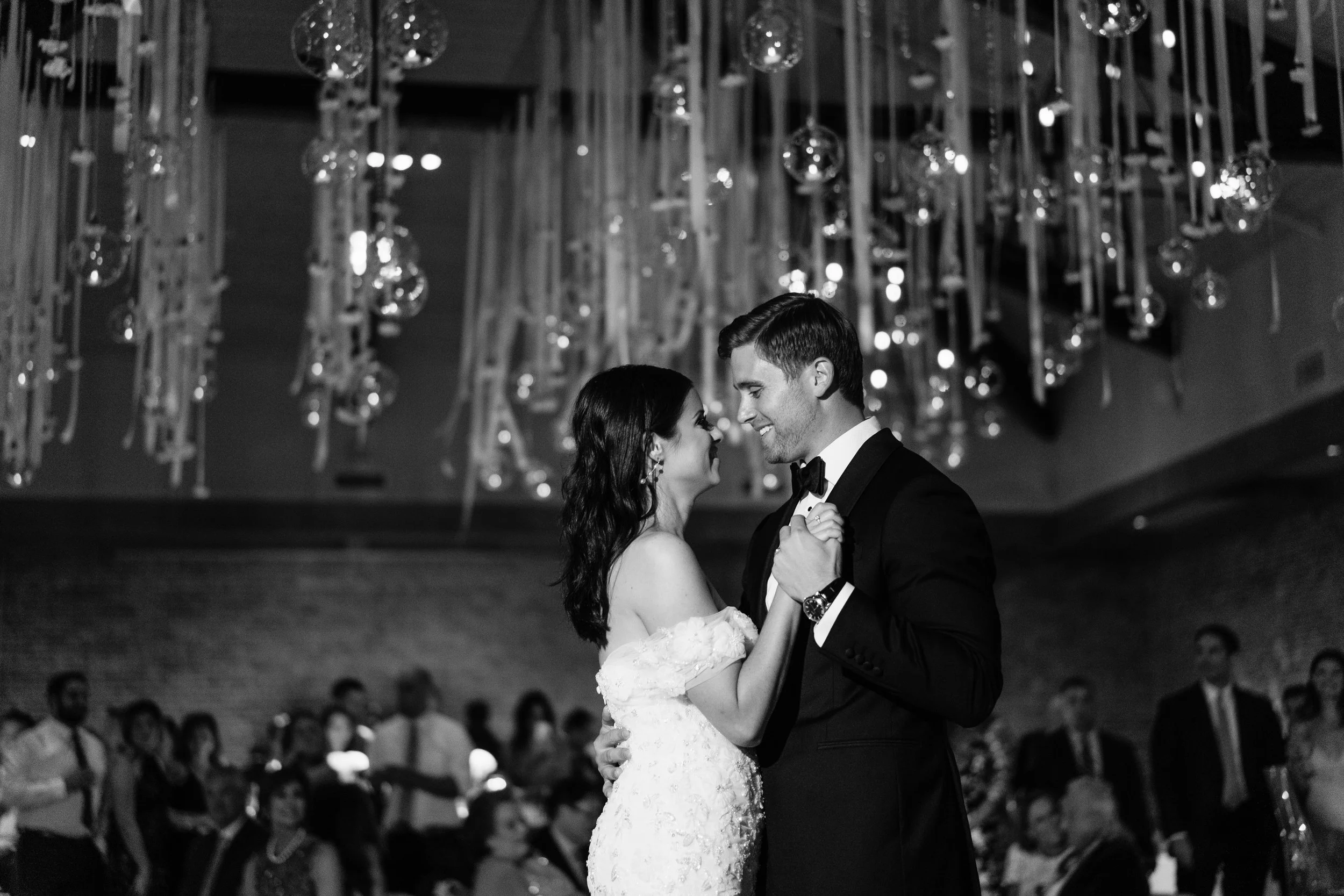 A bride and groom dancing closely during their wedding reception with guests in the background, decorated with hanging ornaments.