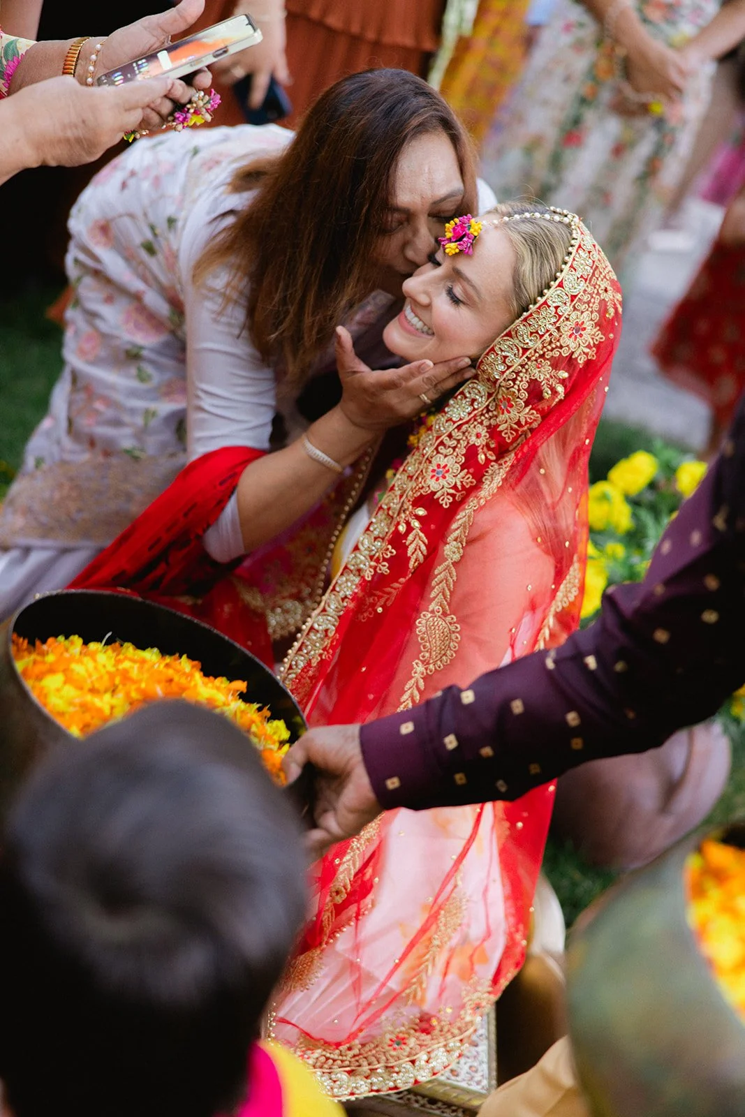 A woman in traditional Indian attire, with a red and gold embroidered saree and a matching head covering, is smiling and embracing an older woman during a cultural or religious ceremony. The women are surrounded by others dressed in colorful clothing