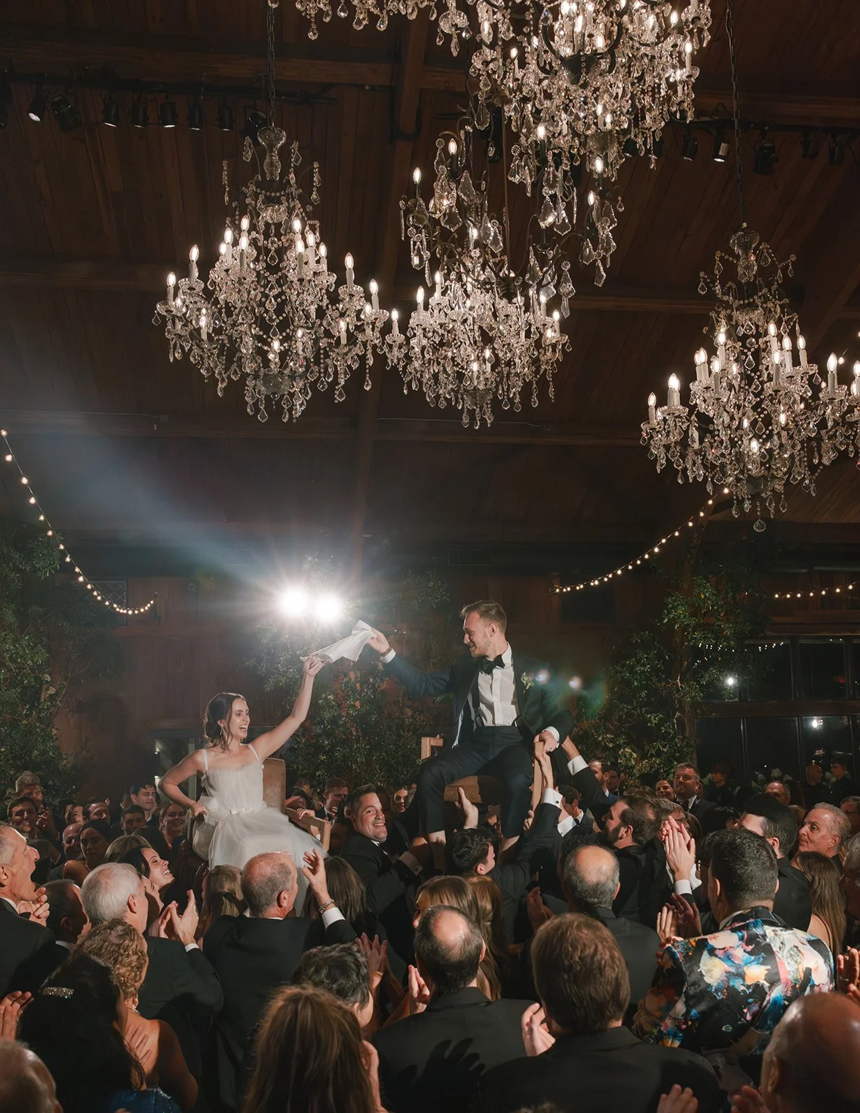 A wedding reception with a bride and groom being lifted on chairs by guests. The bride is in a white dress and the groom is in a suit with a bow tie, smiling and holding a paper. Large chandeliers hang from the ceiling, and the room is decorated with