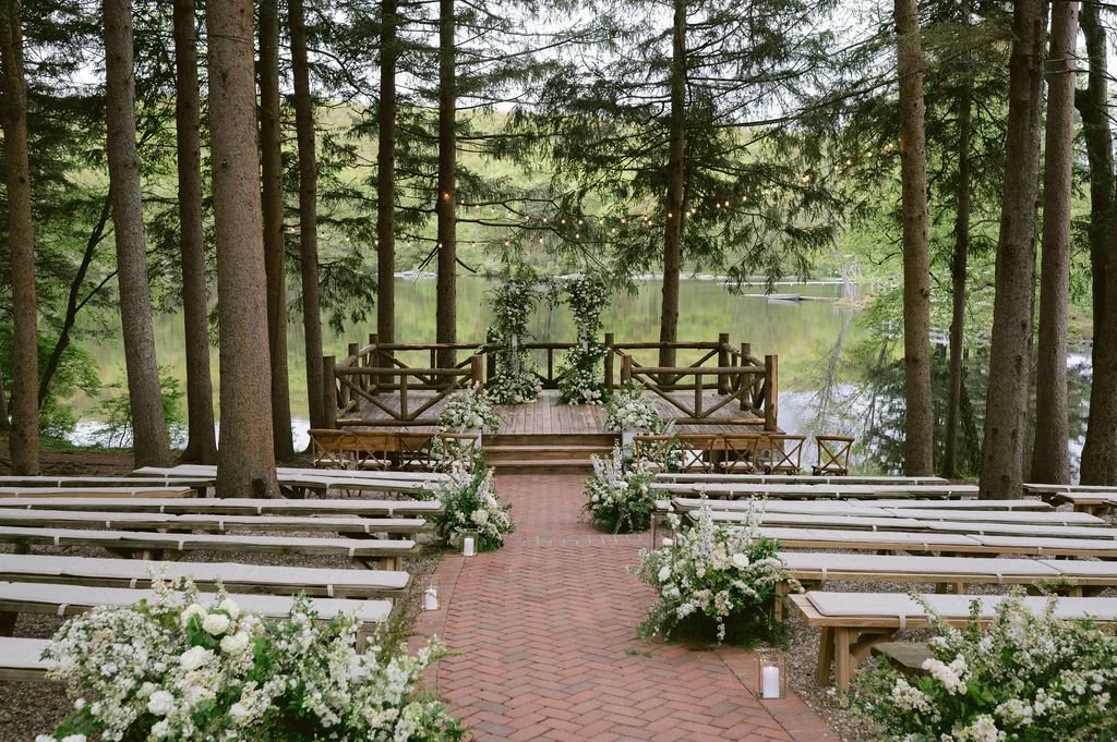 Outdoor wedding setup with white flowers, benches, and a wooden stage decorated with flowers, situated in a wooded area beside a lake.