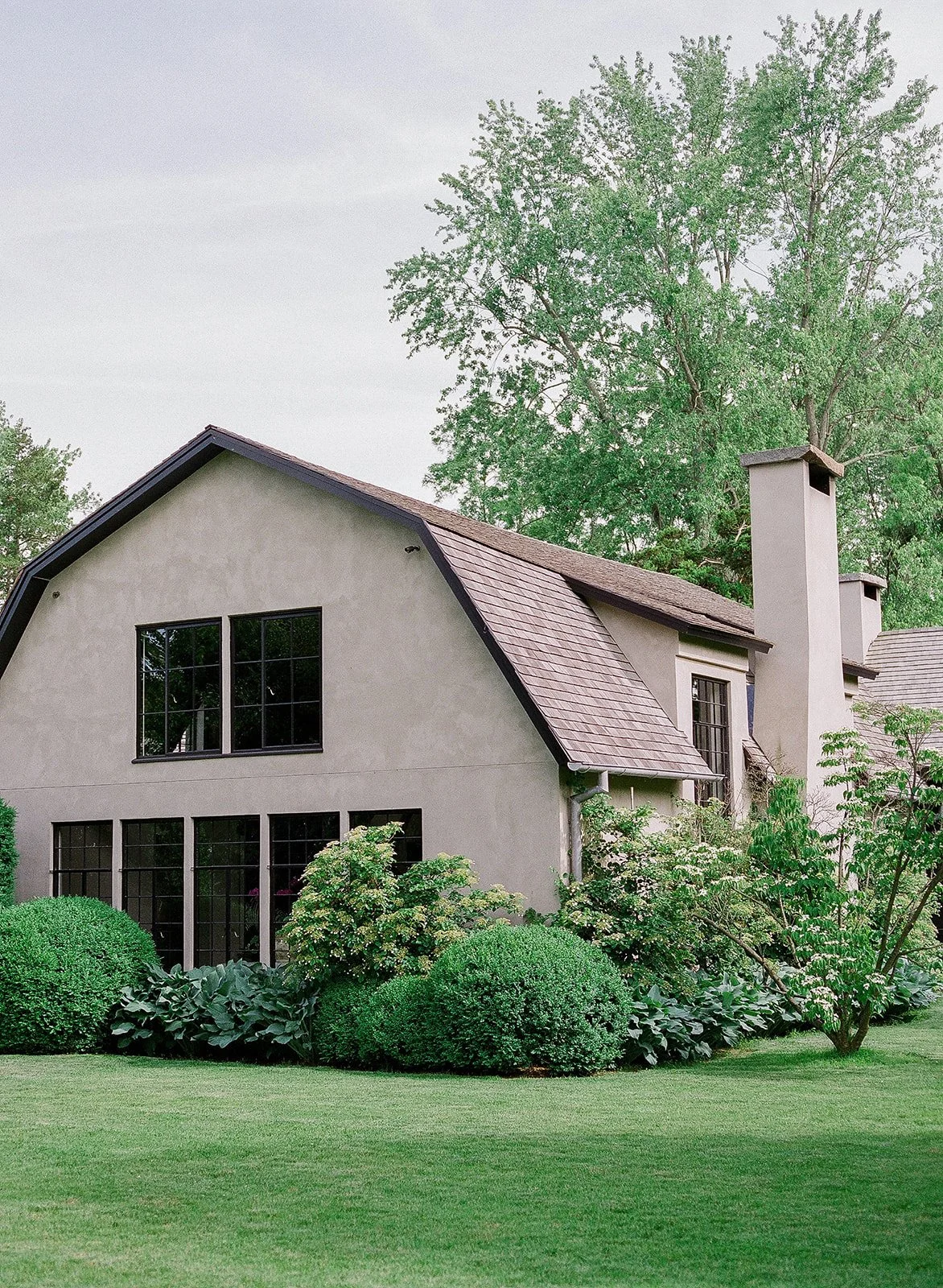 A two-story house with beige stucco exterior, dark window frames, and a gable roof, surrounded by lush green trees and shrubs, with a well-maintained lawn in front.