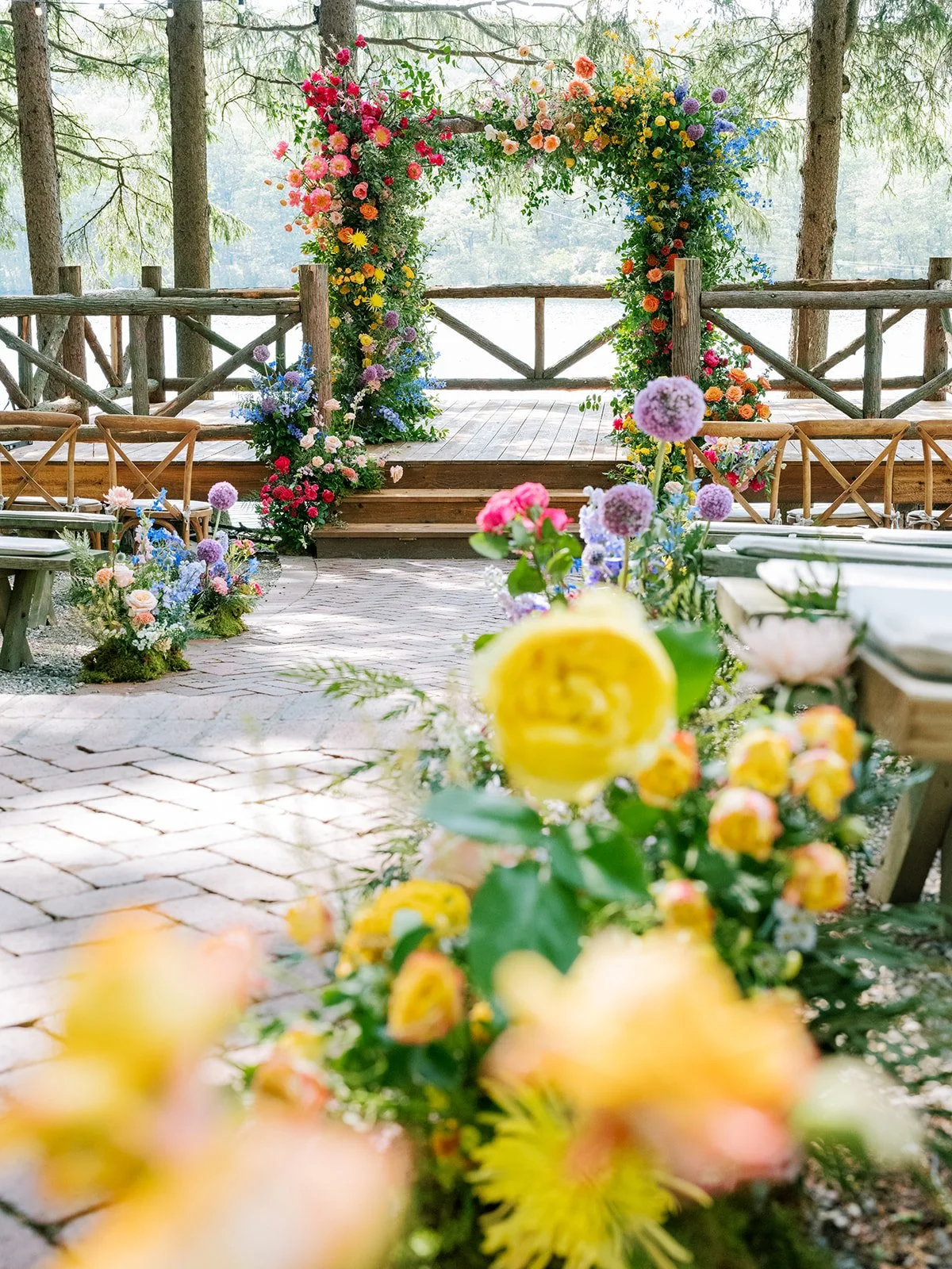 Colorful floral wedding arch with pink, orange, yellow, purple, and blue flowers on a wooden platform in a wooded outdoor setting.