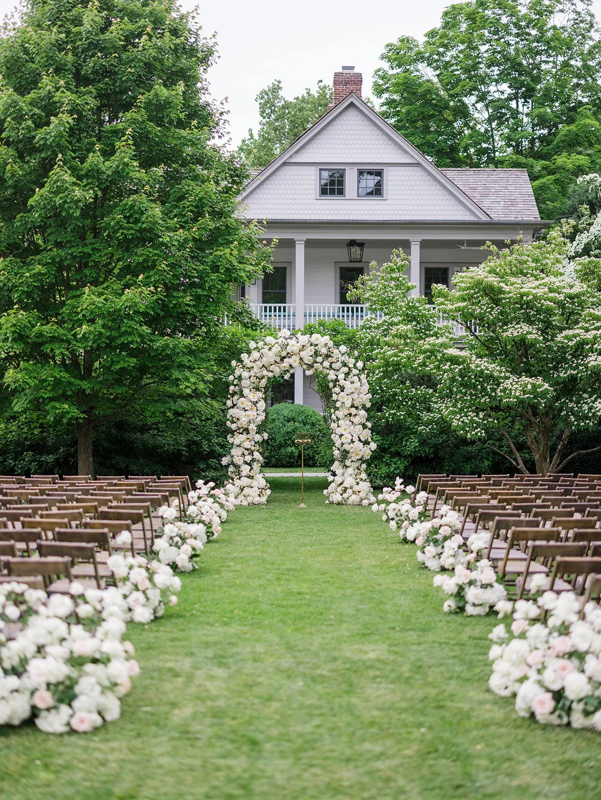 Wedding ceremony setup outdoor with chairs, floral arch, and a house in the background.