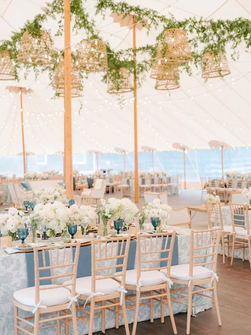Wedding reception decorated with floral centerpieces, string lights, and wicker chandeliers inside a large tent.