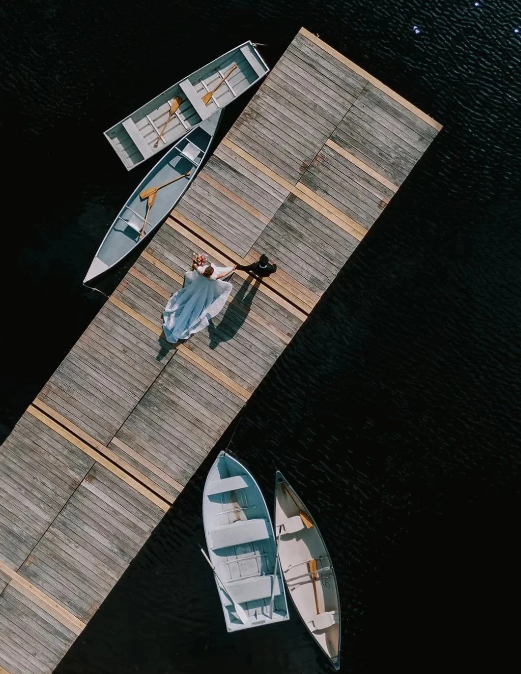 A bride and groom holding hands walking on a wooden dock over water, with two boats docked on either side.