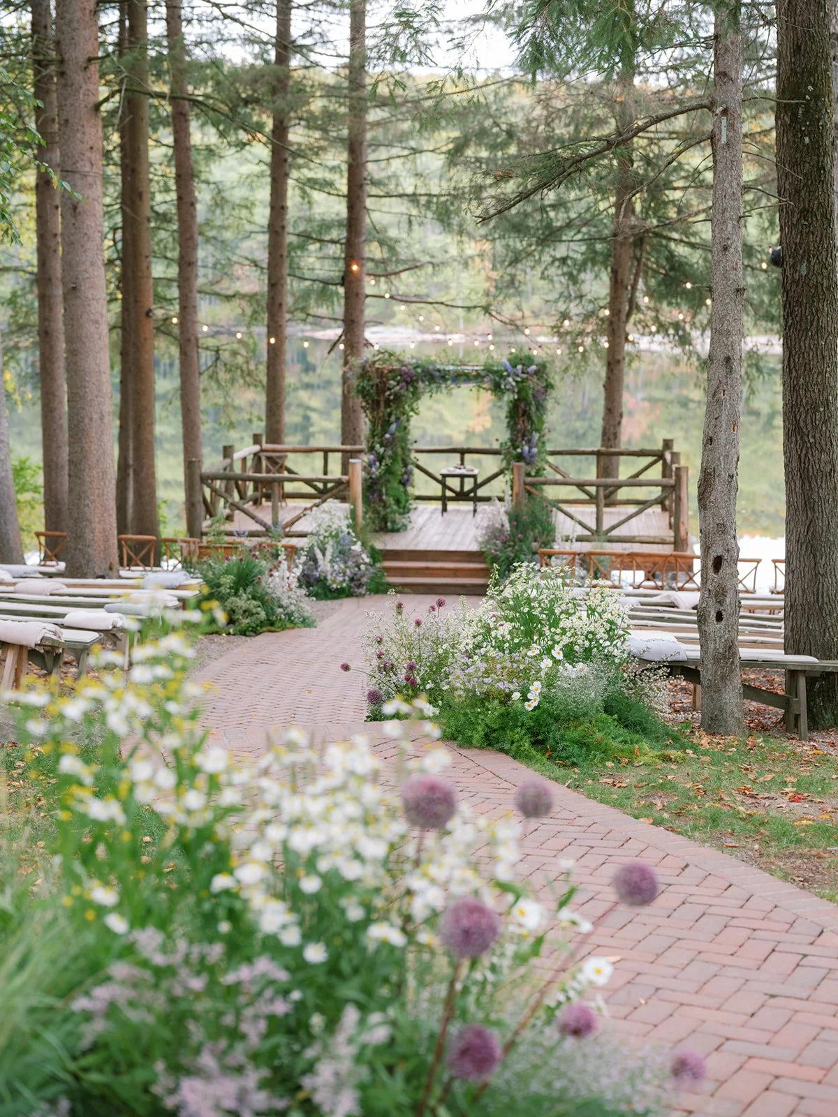 An outdoor wedding ceremony setup in a forested area with a wooden aisle, floral decorations, and a small platform with a flower arch, surrounded by trees and string lights.