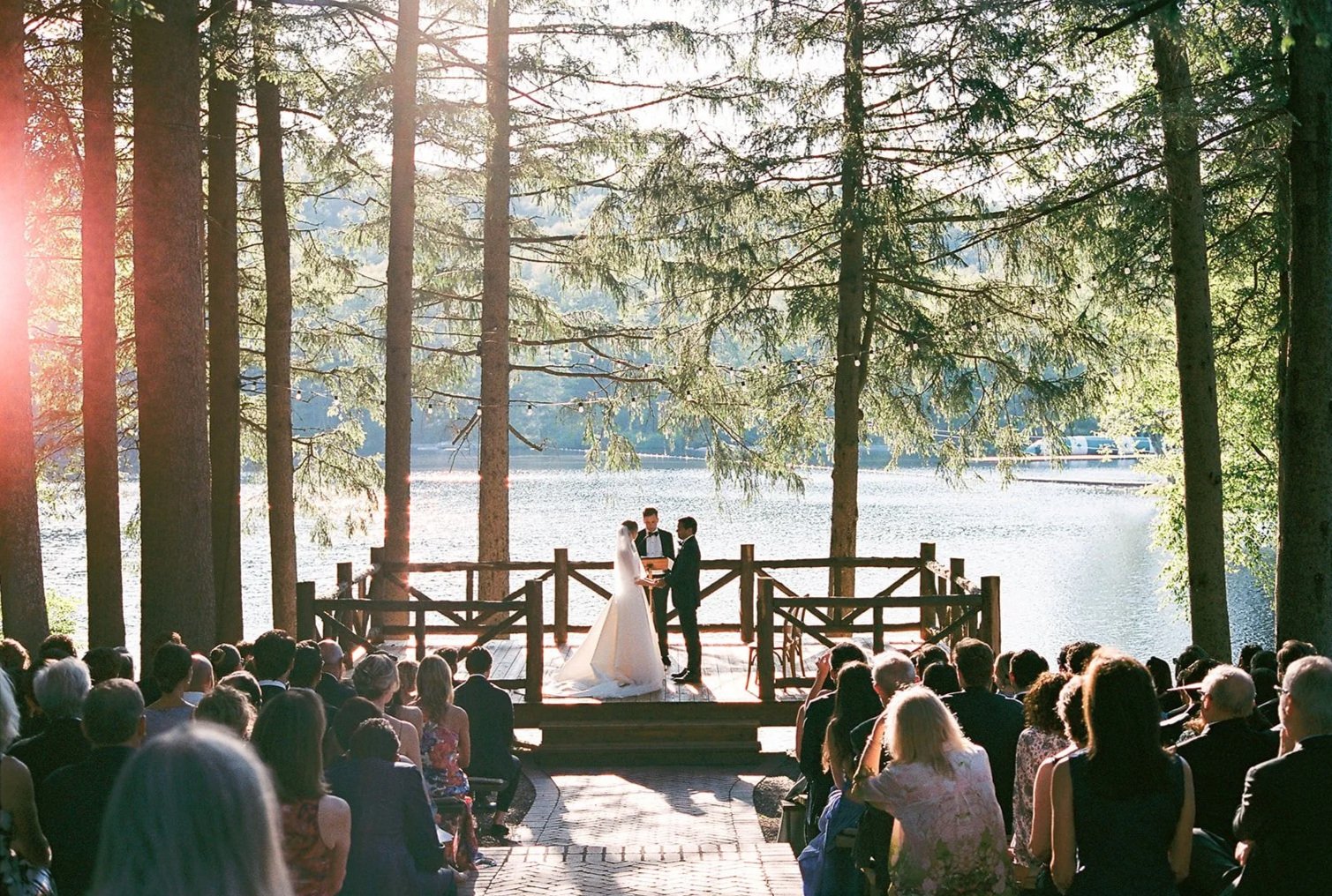 A wedding ceremony outdoors by a lake, with a bride and groom exchanging vows on a wooden platform surrounded by tall trees and toasting guests seated in rows.
