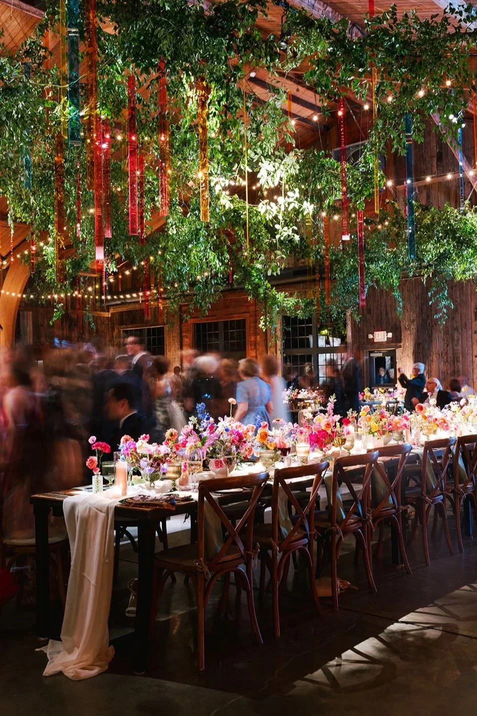 A lively indoor event with a long table decorated with colorful floral arrangements, surrounded by wooden chairs, with people dancing and socializing in a rustic wooden venue decorated with hanging greenery, string lights, and colorful ribbons.