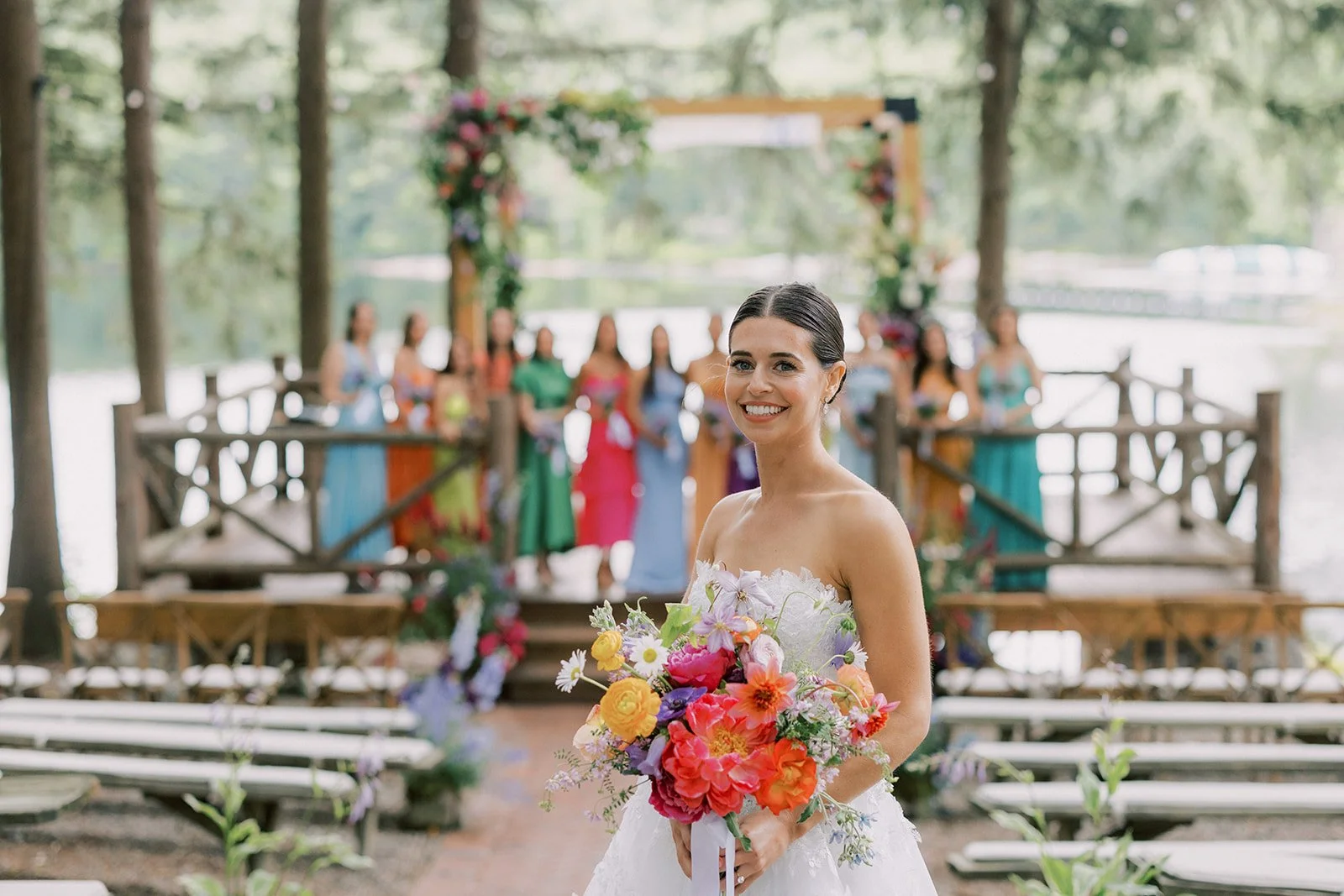 A bride in a white wedding dress holding a bouquet of colorful flowers, smiling in front of a group of women in colorful dresses on a wooden platform outdoors near a lake.