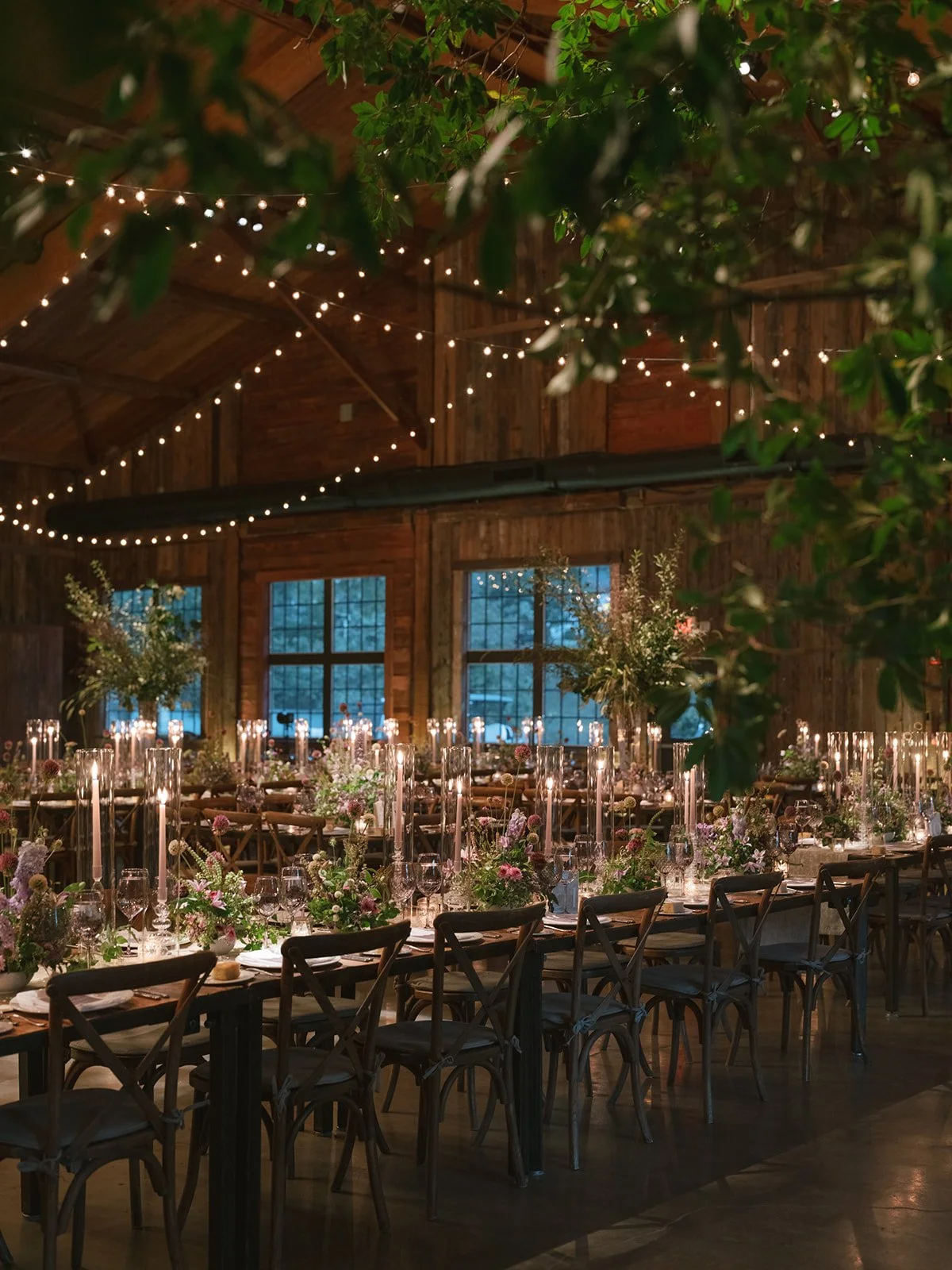 Decorated event hall with long tables, floral centerpieces, candle-like lights, and string lights hanging from the ceiling, in a rustic wooden building.