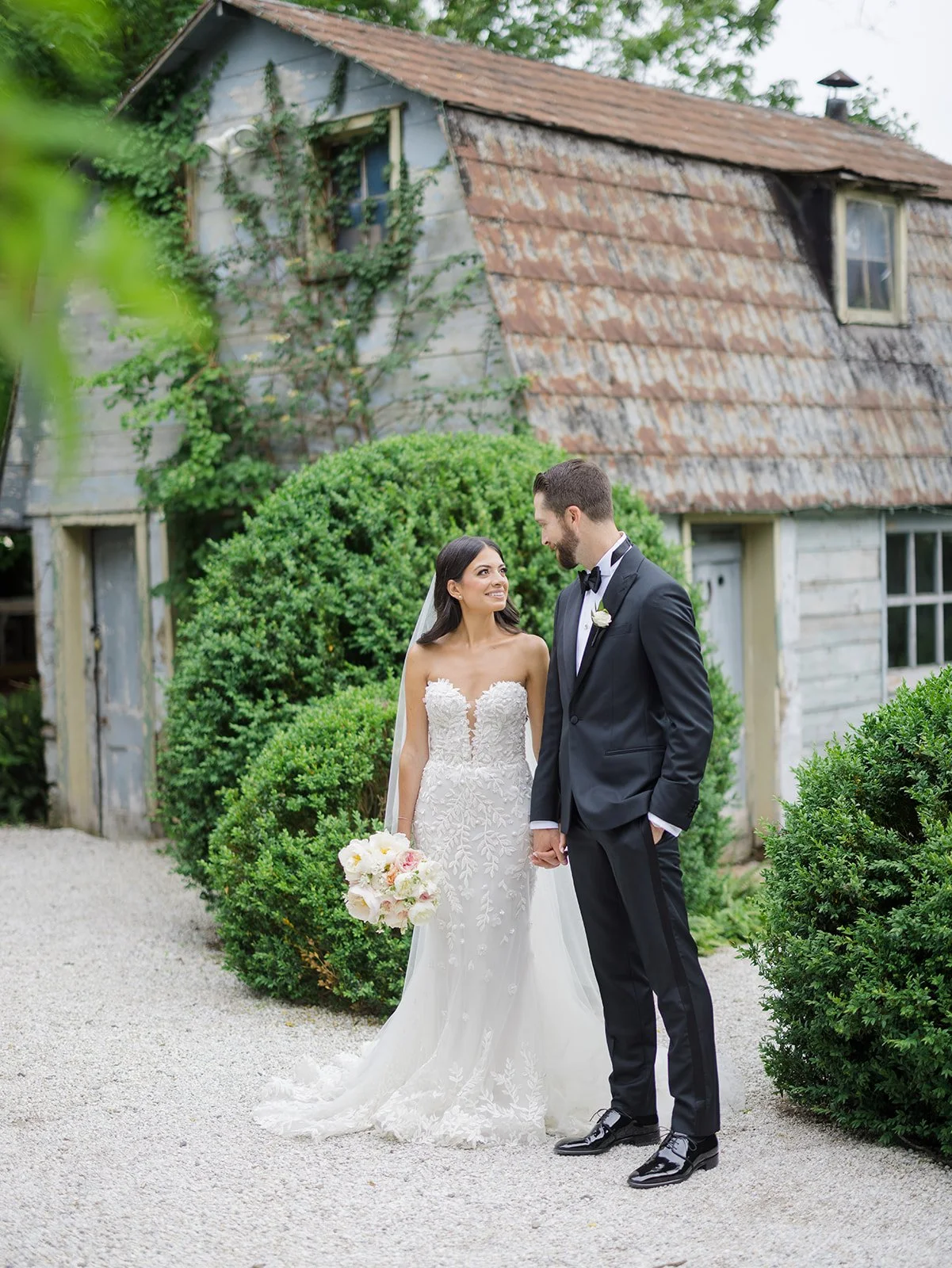 Wedding couple holding hands and smiling at each other outdoors near an old rustic house with greenery.