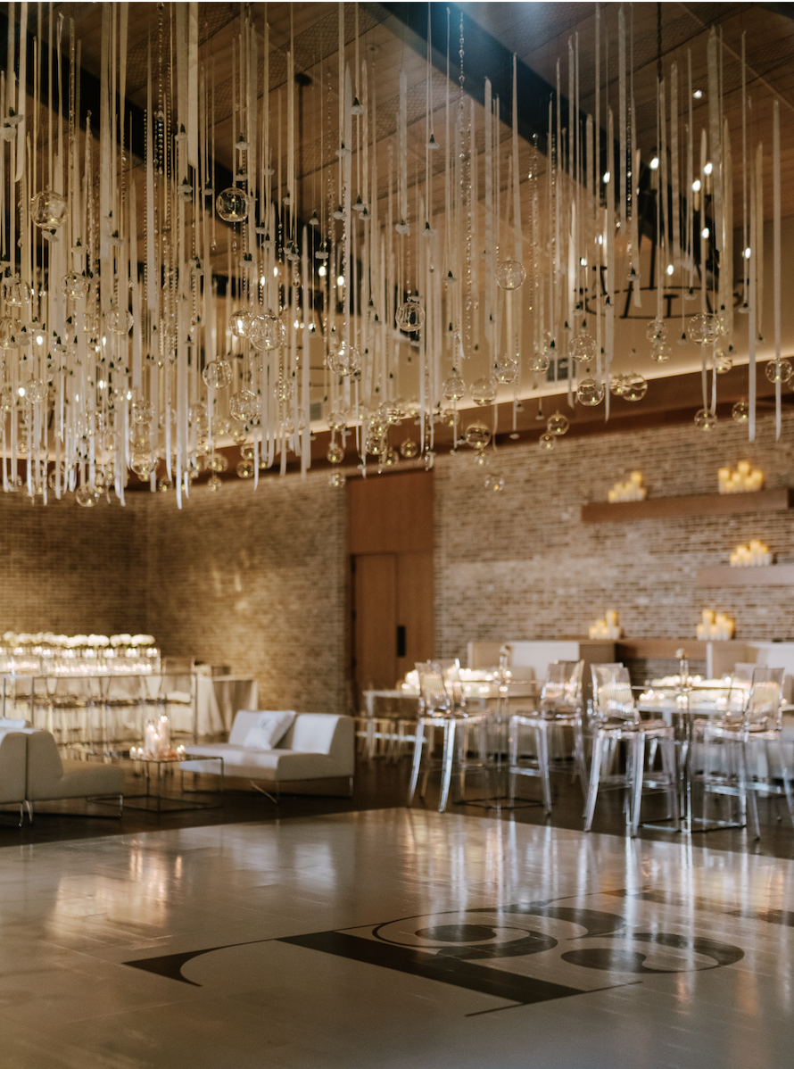 Elegant event space with hanging glass decorations, white sofas, and clear chairs set around tables with candles, illuminated by soft lighting against a brick wall.