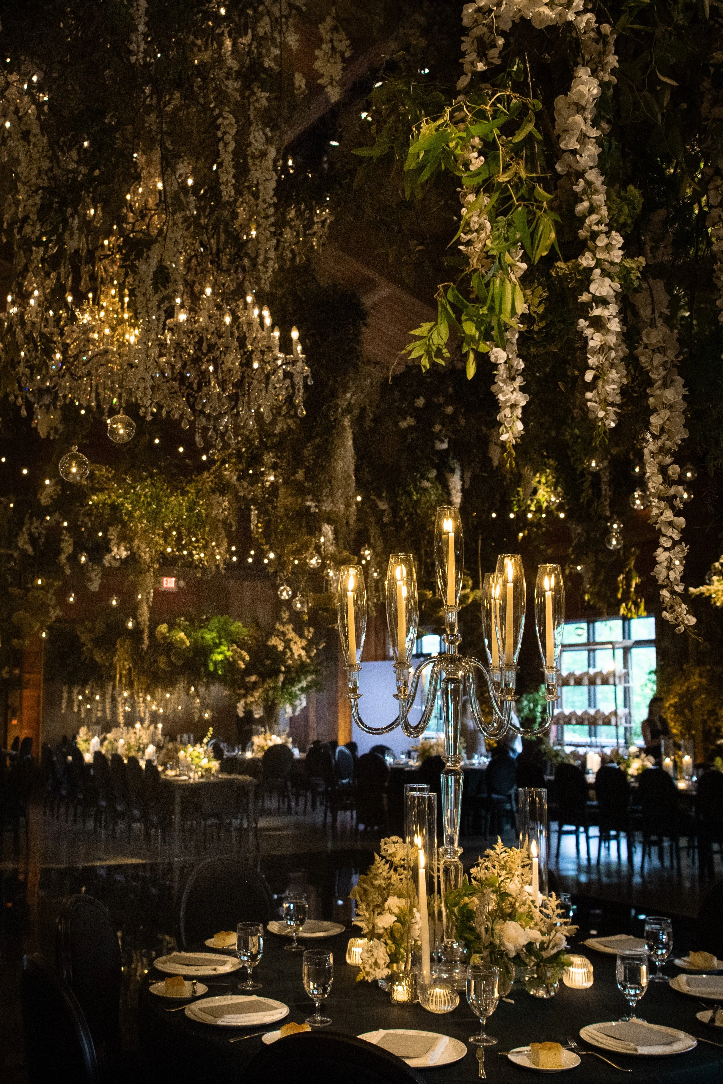 Elegant wedding reception or banquet hall decorated with hanging white flowers and chandeliers, round tables with formal place settings, tall floral centerpieces, and warm ambient lighting.