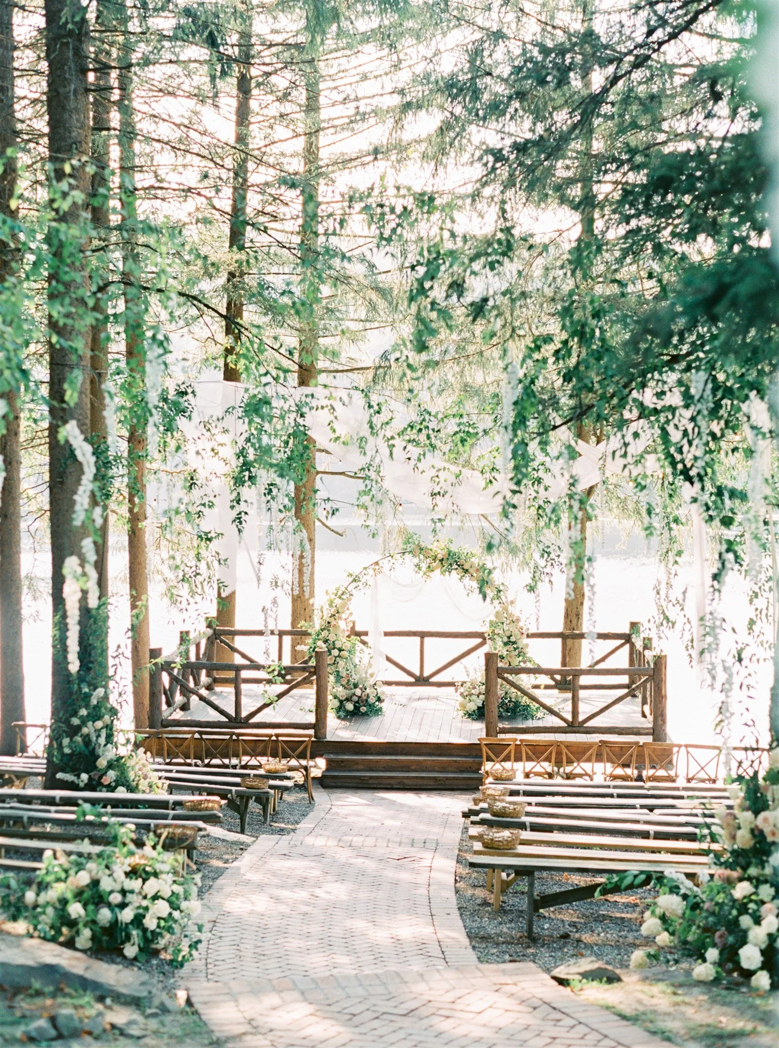 A wedding ceremony setup on a wooden platform in a forest with chairs, floral arrangements, and an arched floral decoration.