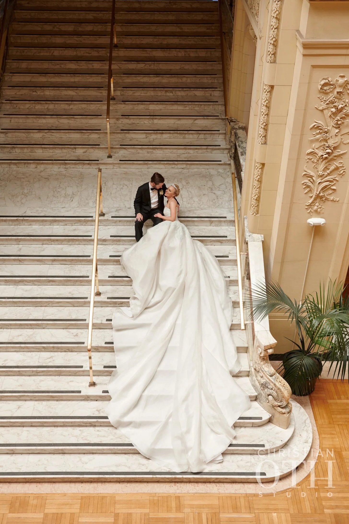 Bride and groom seated on marble staircase, wedding dress has a long train, ornate architecture and a potted plant nearby.