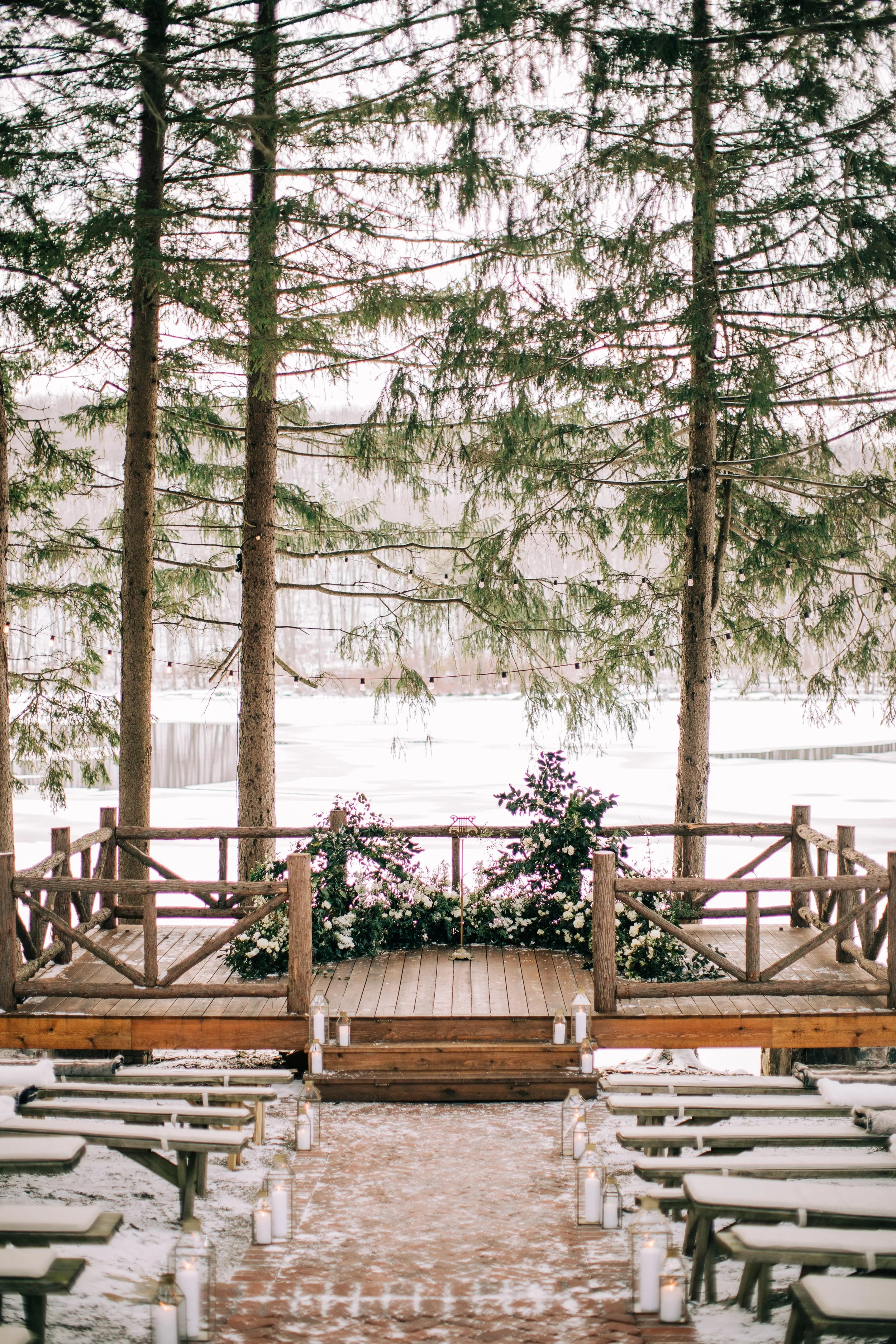 Outdoor wedding ceremony setup on a wooden deck in a snowy forest, decorated with flowers and candles.