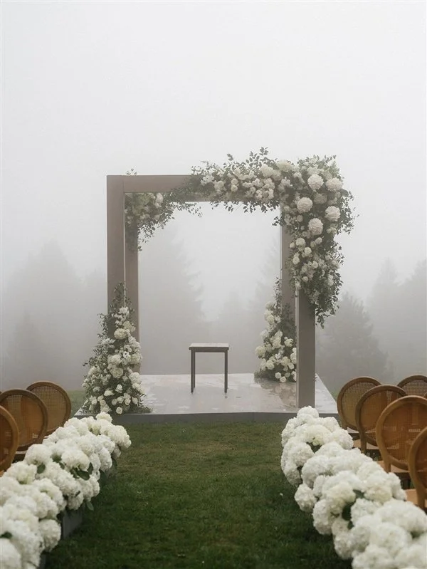 Empty wedding altar decorated with white flowers and greenery, set outdoors on a foggy day with chairs lined up on each side.