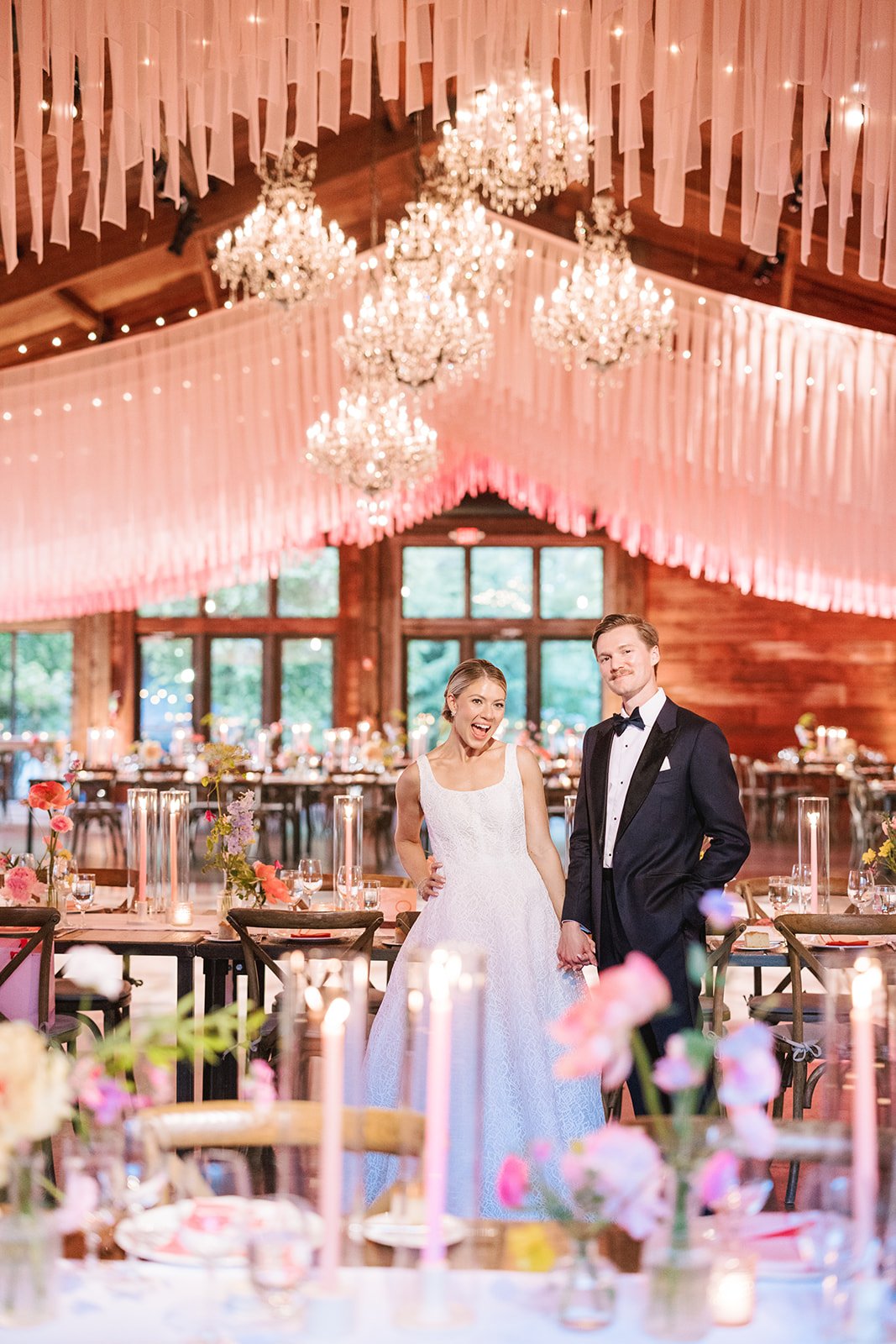 Bride and groom in wedding attire standing inside a decorated reception hall with pink drapery, chandeliers, and tables with flowers and candles.