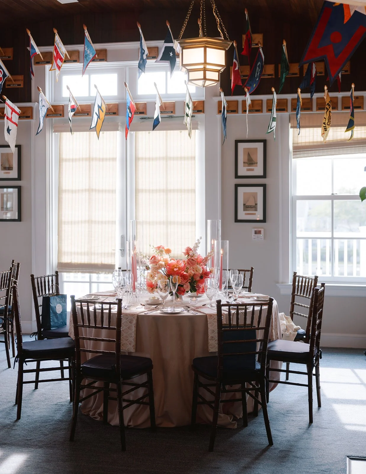 A round dining table with a floral centerpiece and pink candles, set for a formal event, in front of large windows with beige blinds, surrounded by dark wooden chairs, decorated with nautical flags hanging from the ceiling.