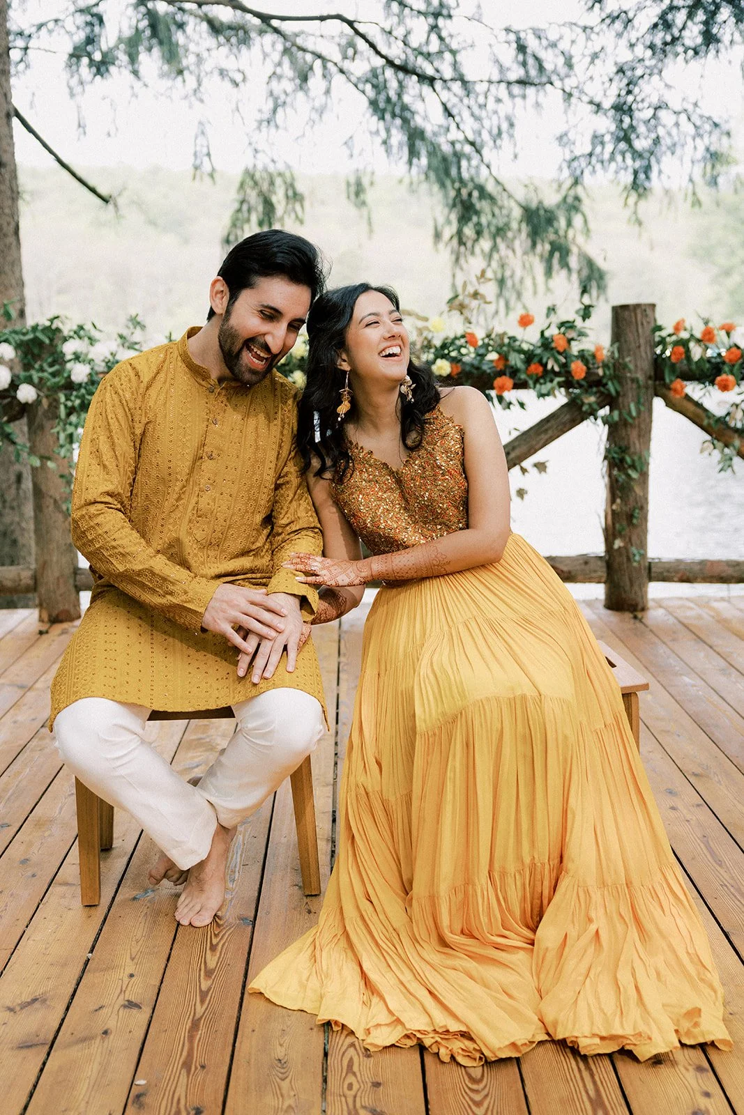 A man and woman sitting on a wooden deck, dressed in traditional Indian attire, laughing and enjoying each other's company with a lakeside backdrop and floral decorations.