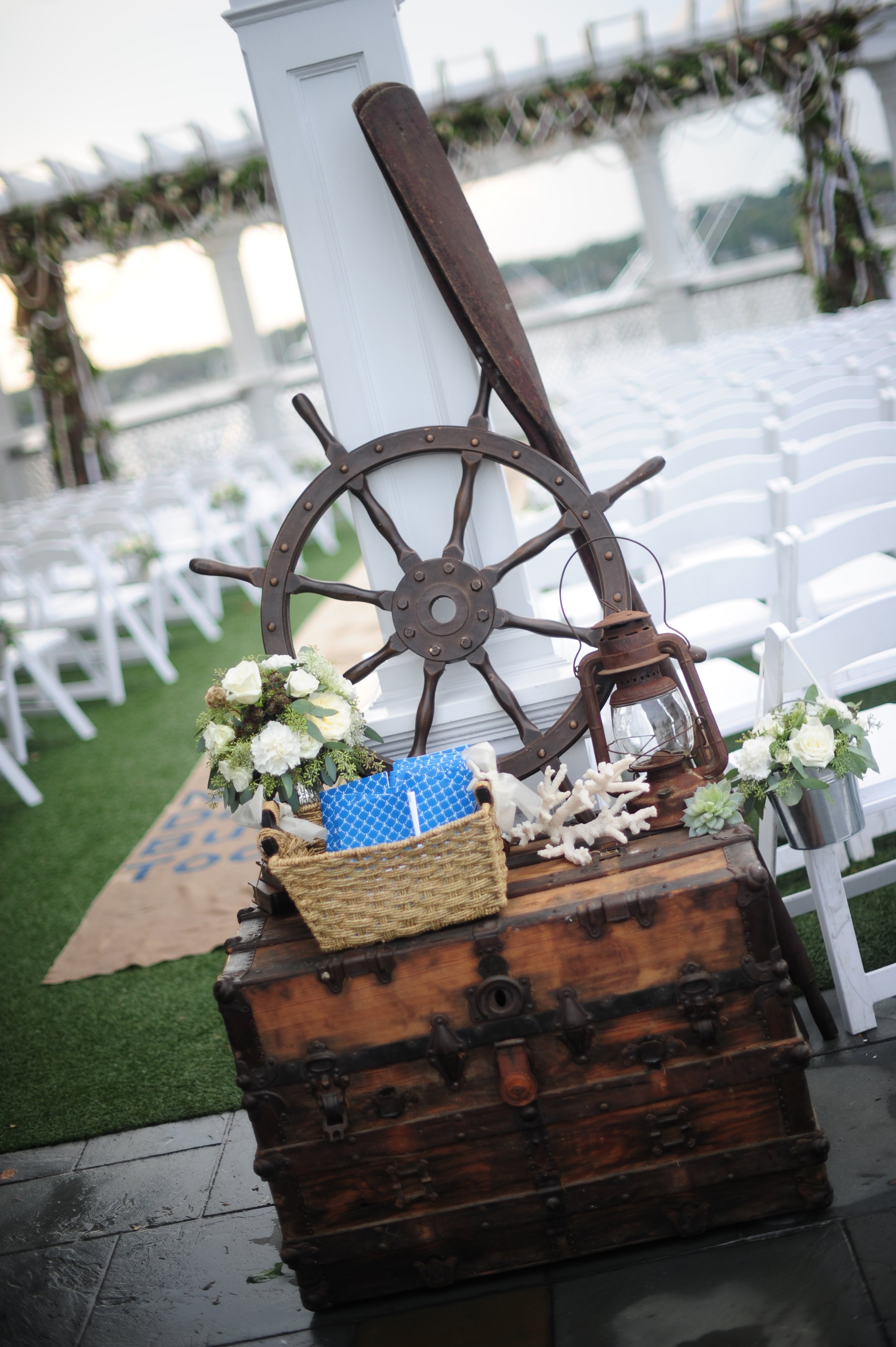 Decorative nautical-themed wedding setup on a dock with white chairs in the background, featuring a wooden chest, steering wheel, lantern, flowers, and coral decoration.