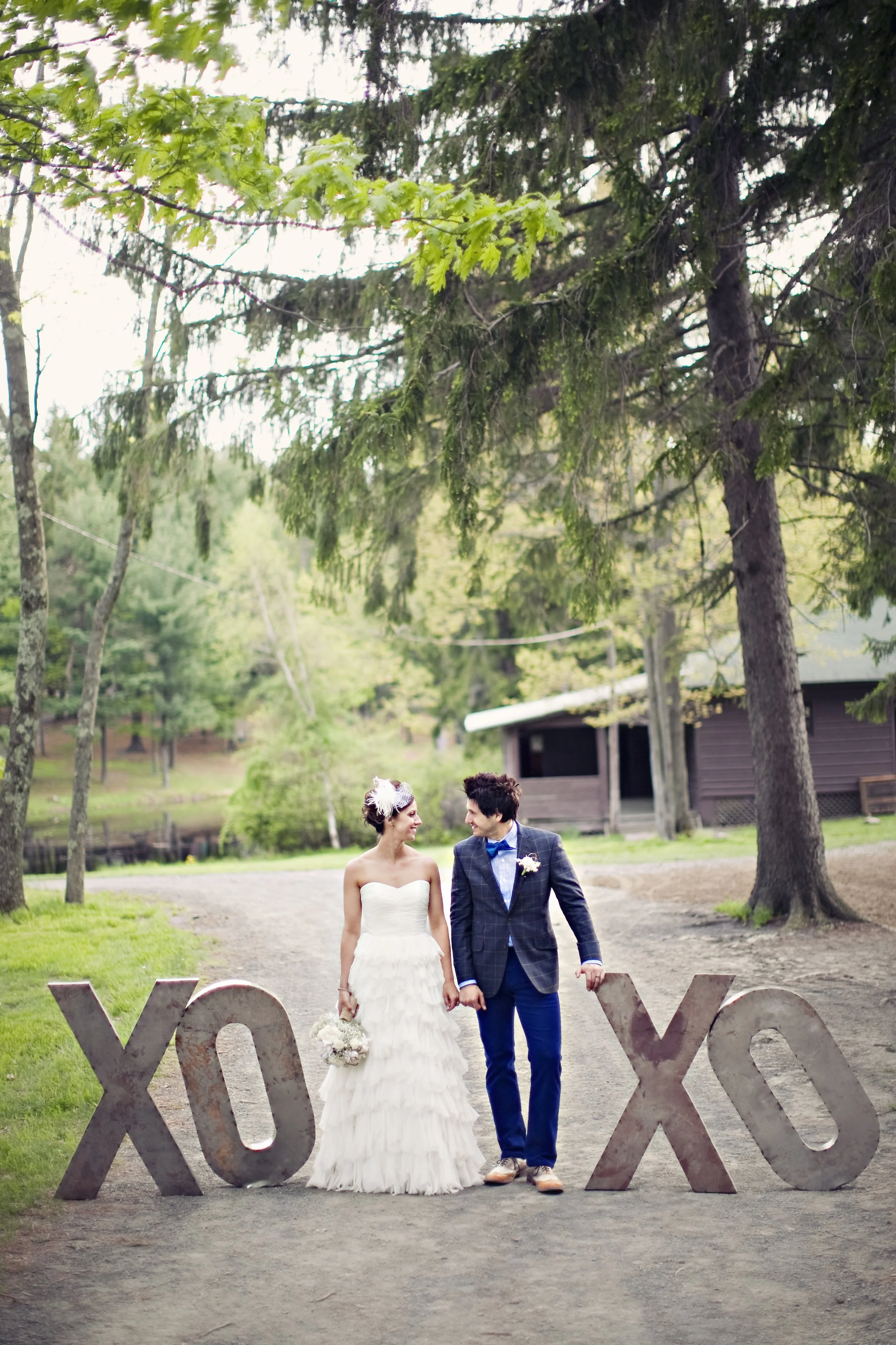 A bride and groom holding hands, standing outdoors with large XO decorations around them, surrounded by trees and a cabin in the background.