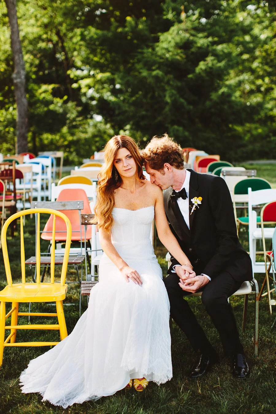 A bride and groom sitting outdoors on a grassy area with colorful chairs in the background, holding hands and sharing a tender moment.
