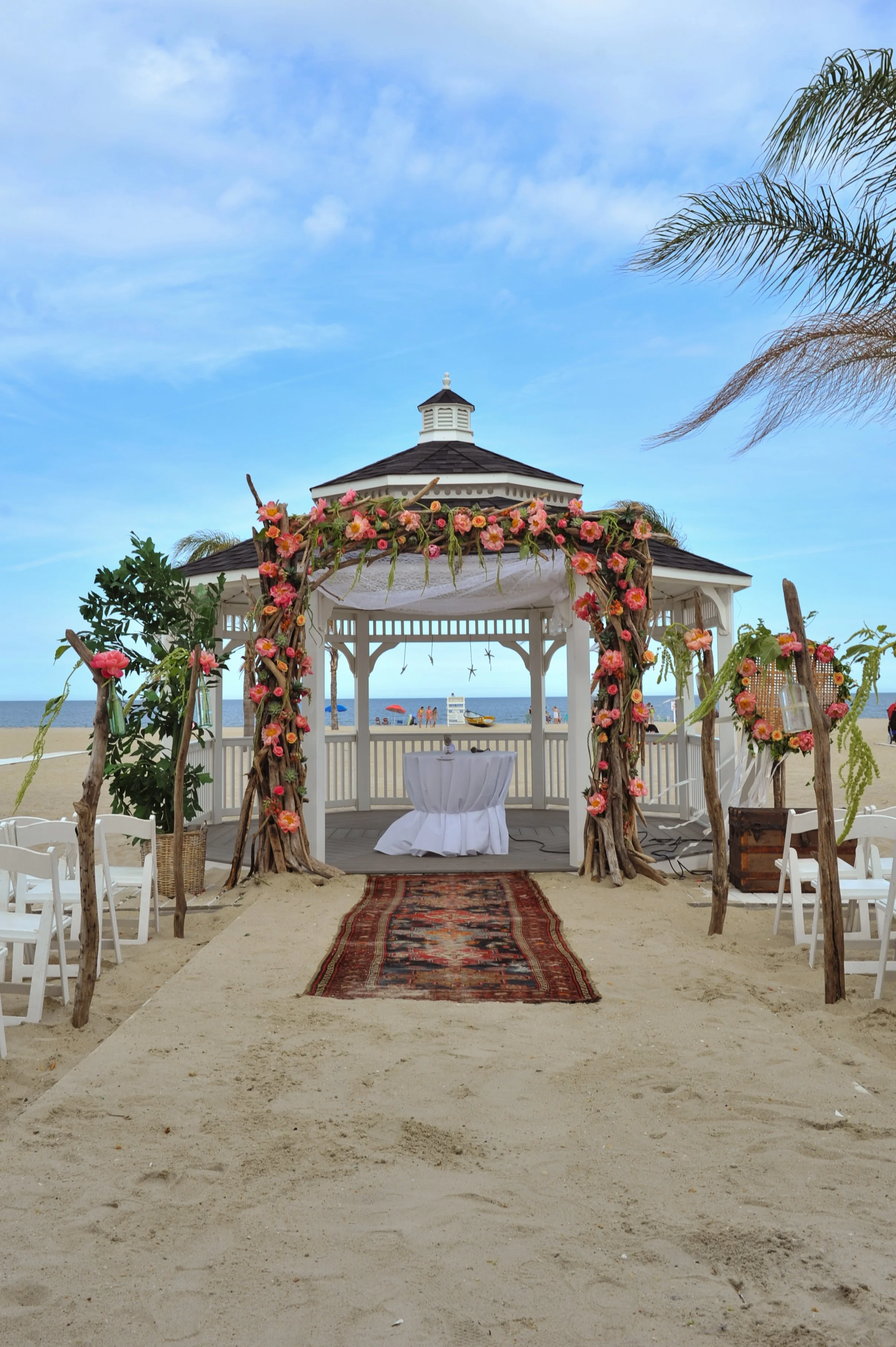 Beach wedding setup with a gazebo decorated with pink flowers, a table with a white tablecloth, surrounded by white chairs, and a sandy pathway leading to the ocean under a blue sky.