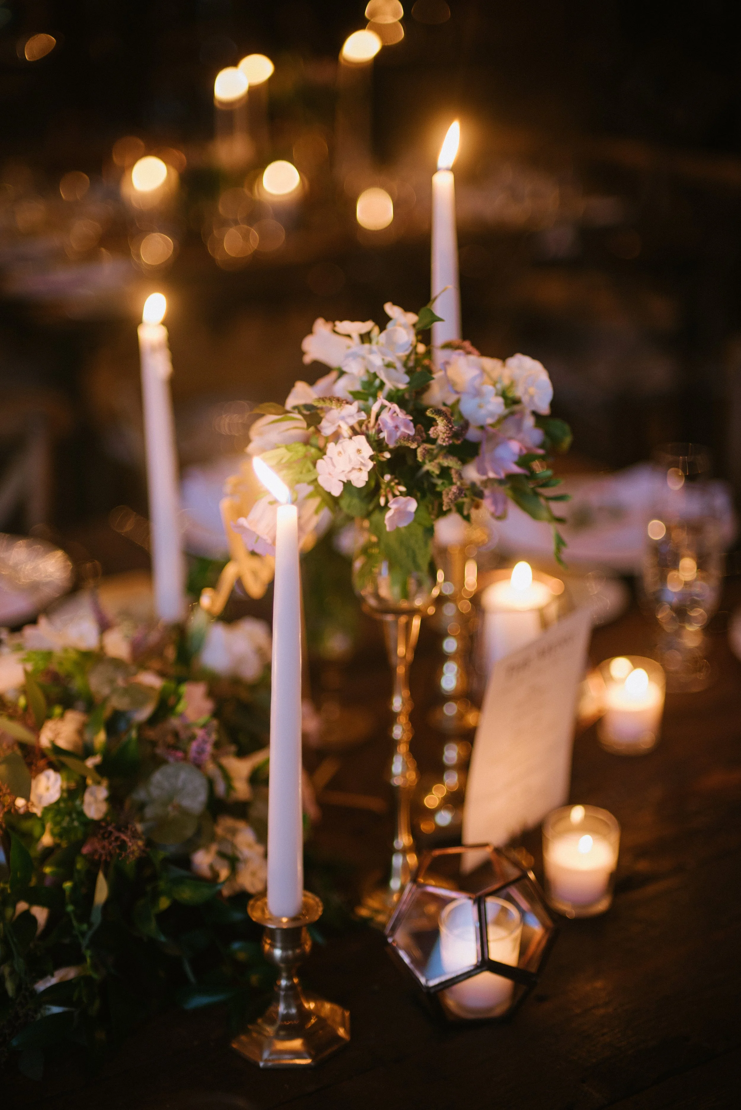 A decorated dinner table with lit white candles, floral arrangements, and small votive candles in a warm, dimly lit setting.