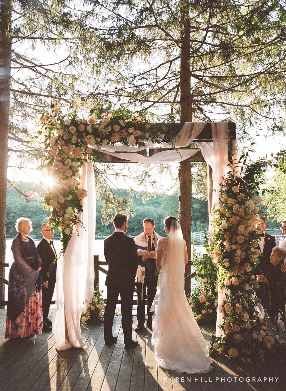 A wedding ceremony taking place outdoors under a floral arch with the bride and groom exchanging vows, surrounded by family and friends, during sunset.