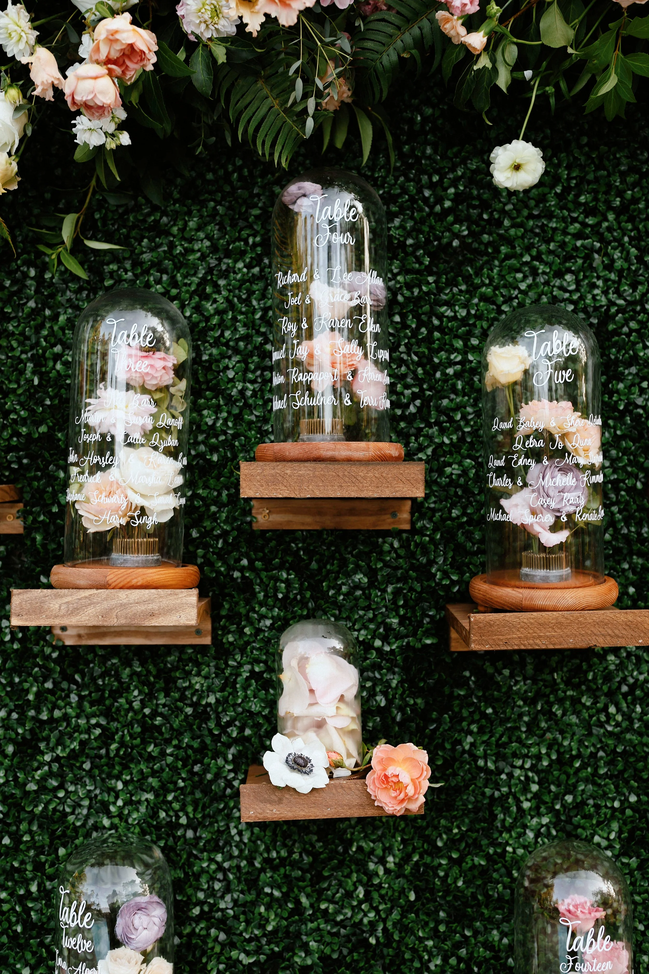Decorative wall display with artificial flowers and glass cloches containing flowers, each labeled with table numbers and guest names for a wedding or event.