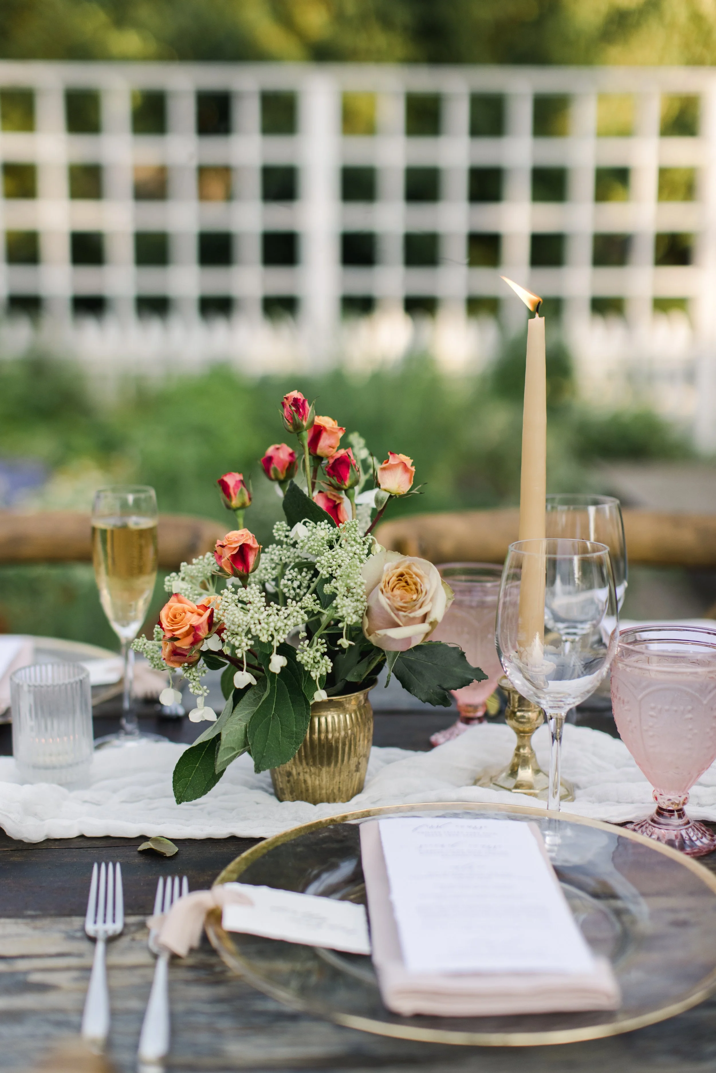 Table setting with a floral arrangement, a tall candle, wine glasses, water glasses, pink goblet, and a menu on a dark wooden table, outdoors with a blurred white fence and greenery in the background.