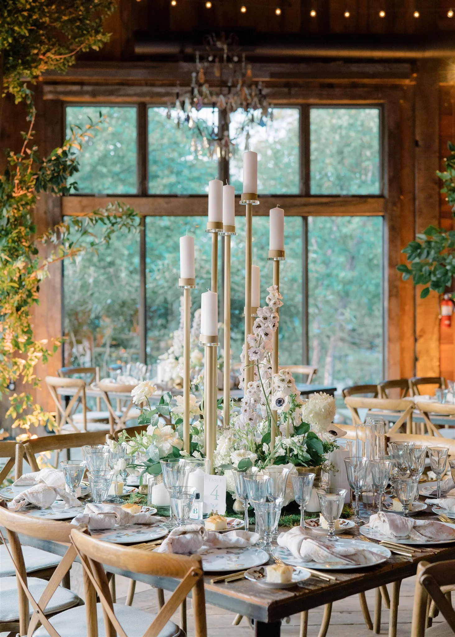 Decorated round dining table set for a formal event in a rustic venue with large windows, floral arrangements, tall candles, glassware, plates, and napkins.