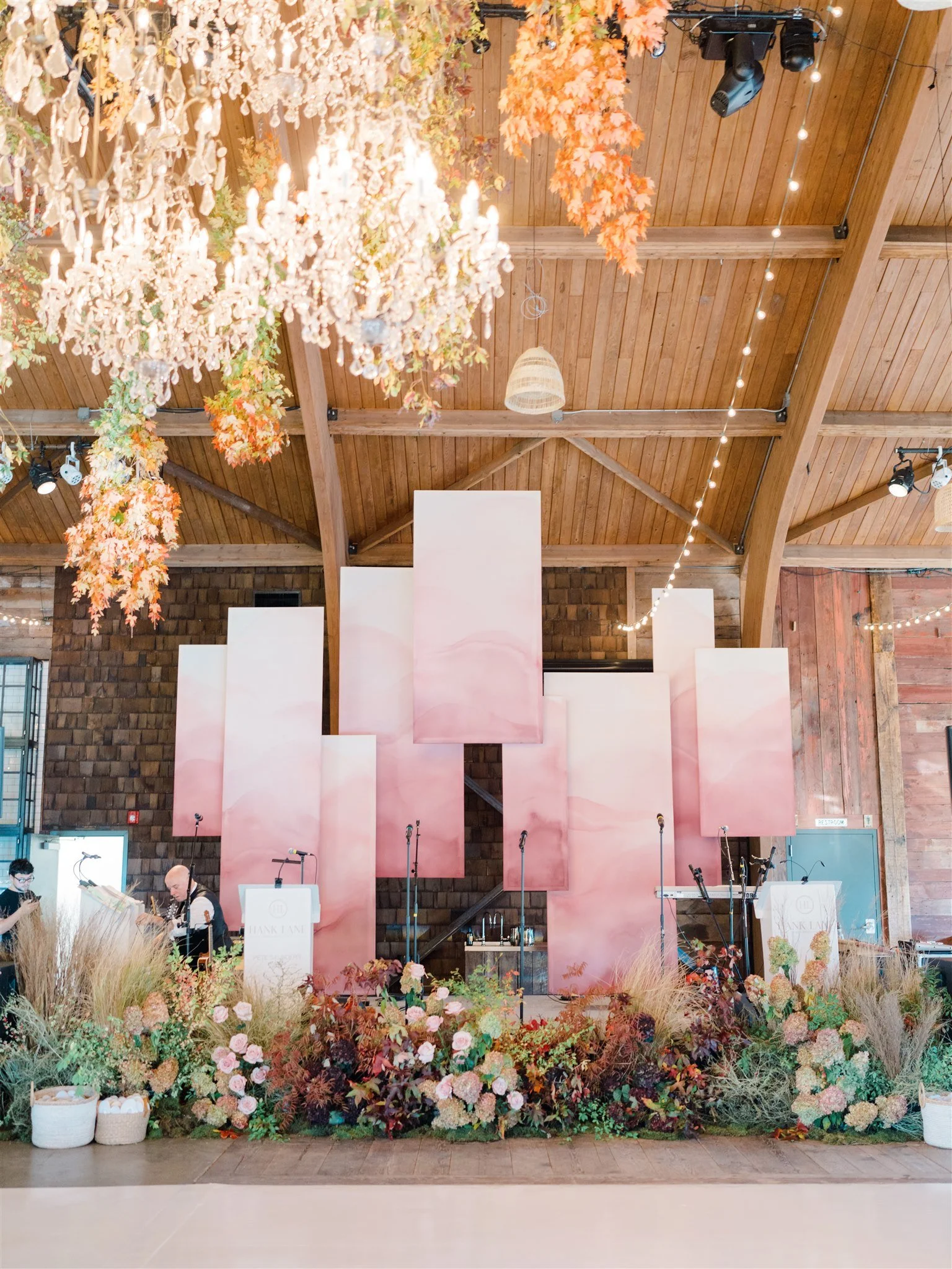 Wedding or event stage decorated with pink and white clouds, floral arrangements, and hanging chandeliers in a wooden barn setting.