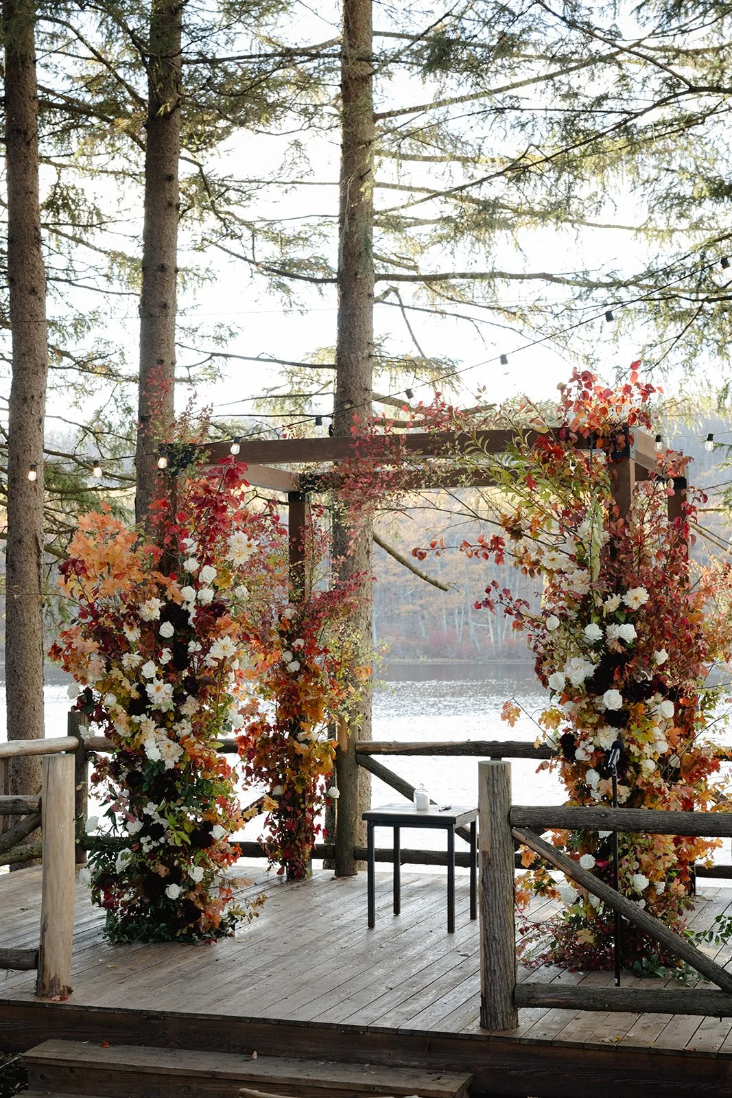 A wooden outdoor wedding altar decorated with floral arrangements of orange, red, and white flowers, set against a backdrop of trees and a lake.