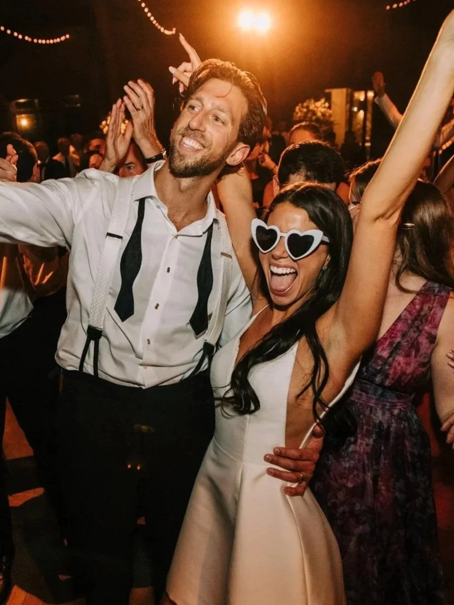 A joyful man and woman dancing at a lively party or celebration, with the woman wearing heart-shaped sunglasses and raising her arm.