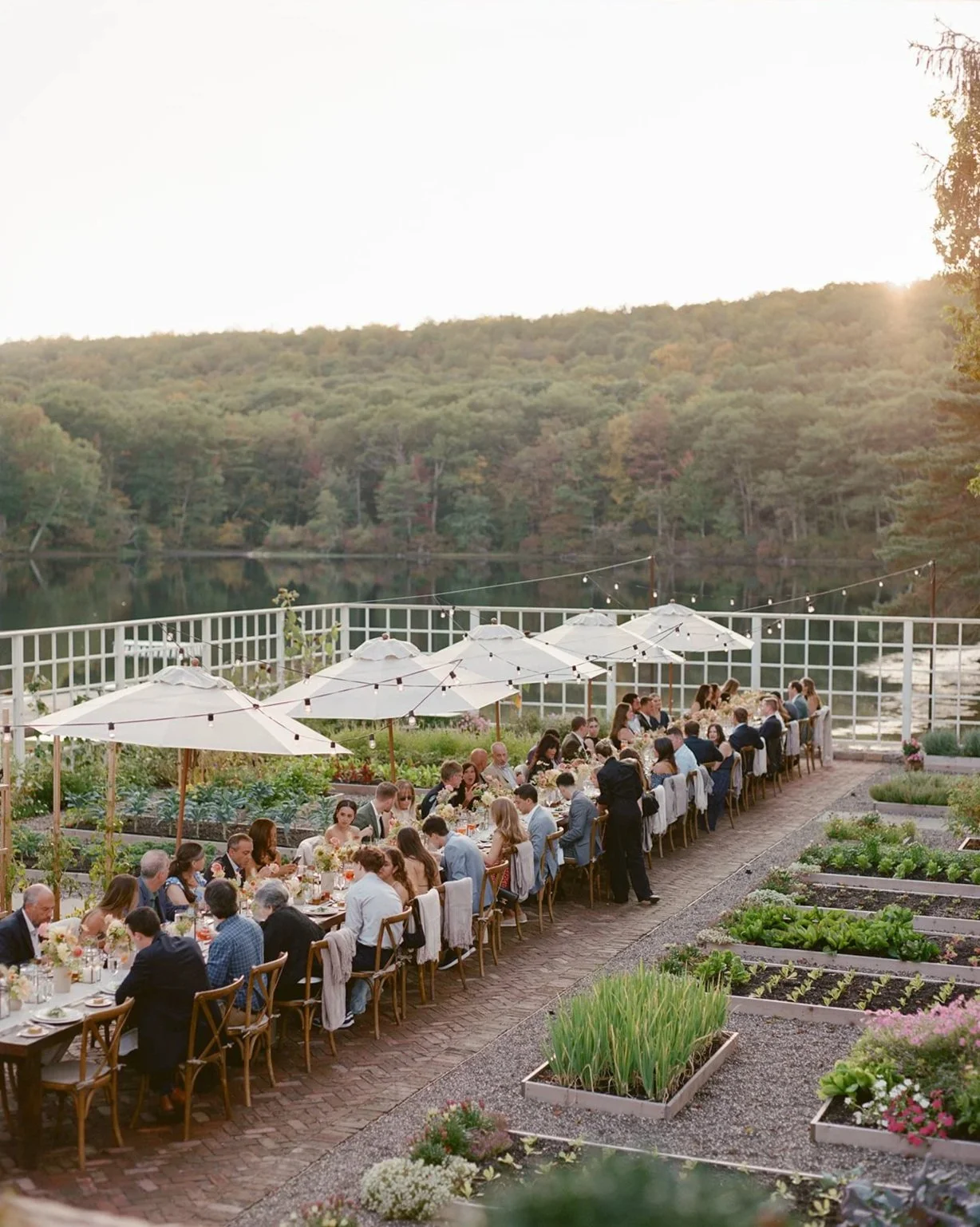 People dining at a long table outdoors with umbrellas, set in a garden with vegetable beds, overlooking a lake and hills at sunset.