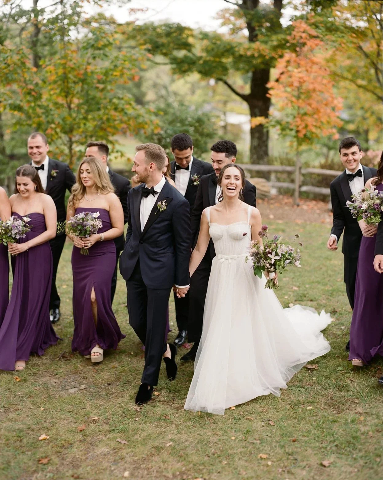 Bride and groom holding hands and walking outdoors with wedding party, surrounded by greenery and autumn trees.