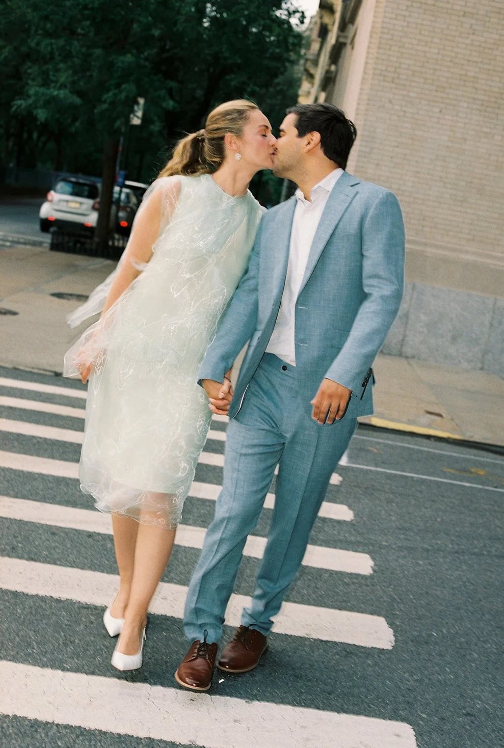 A bride and groom share a kiss and hold hands while walking across a crosswalk on a city street, dressed in wedding attire.