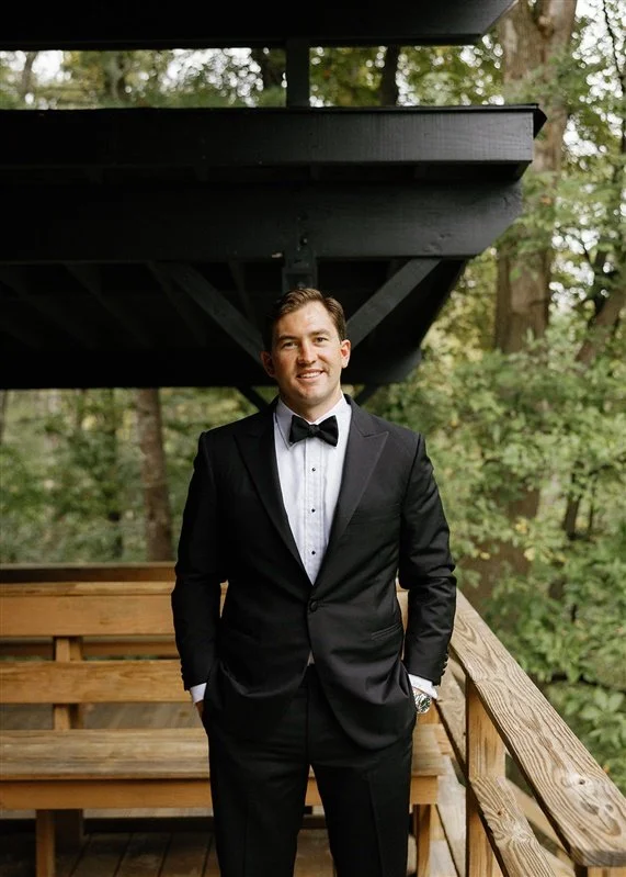 A man in a black tuxedo with a white shirt and black bow tie standing outdoors on a wooden deck surrounded by green trees.