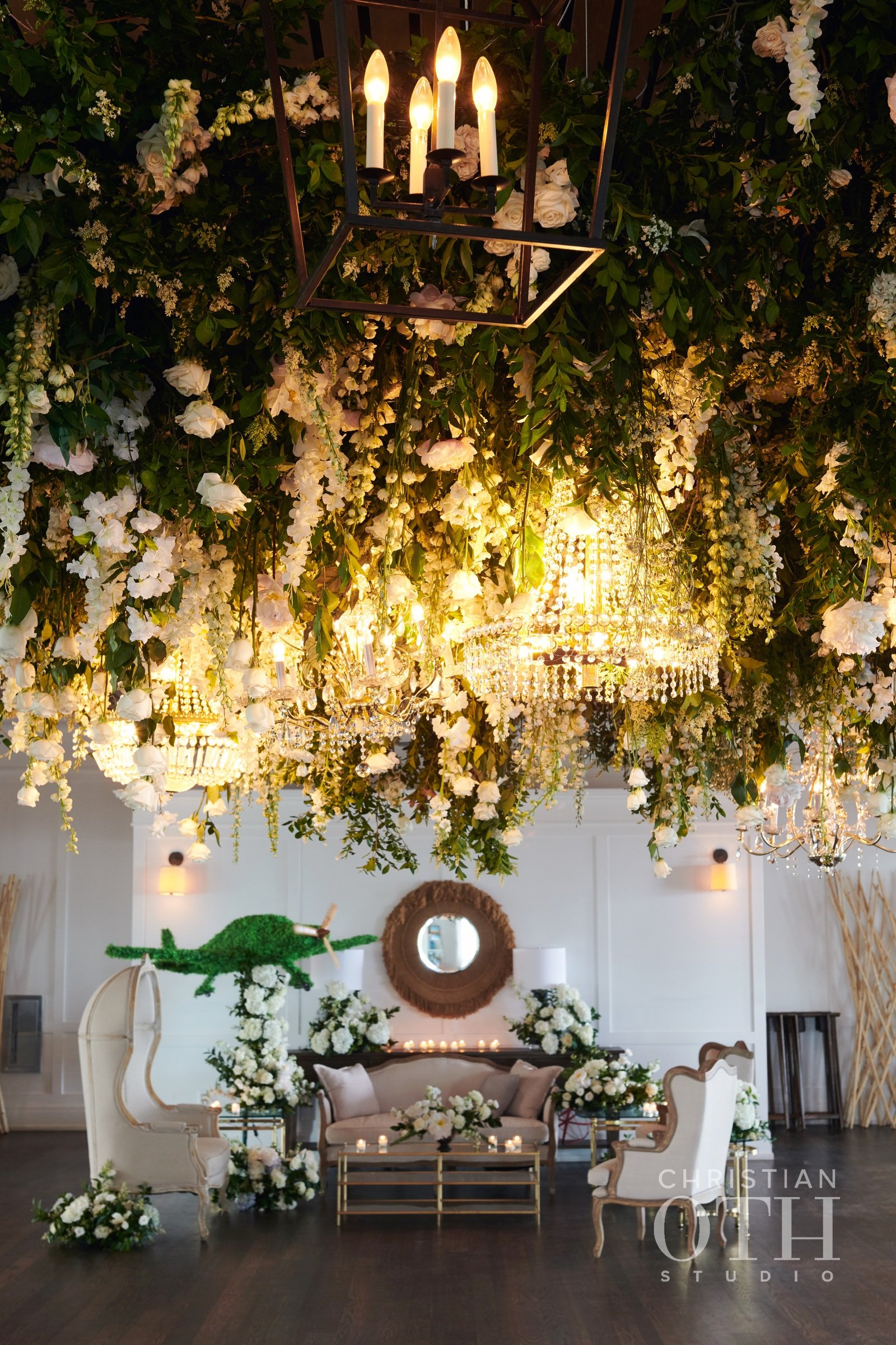 Elegant wedding or event space decorated with a large floral arrangement hanging from the ceiling, chandeliers, and soft lighting, with seating furniture and floral arrangements on the floor.
